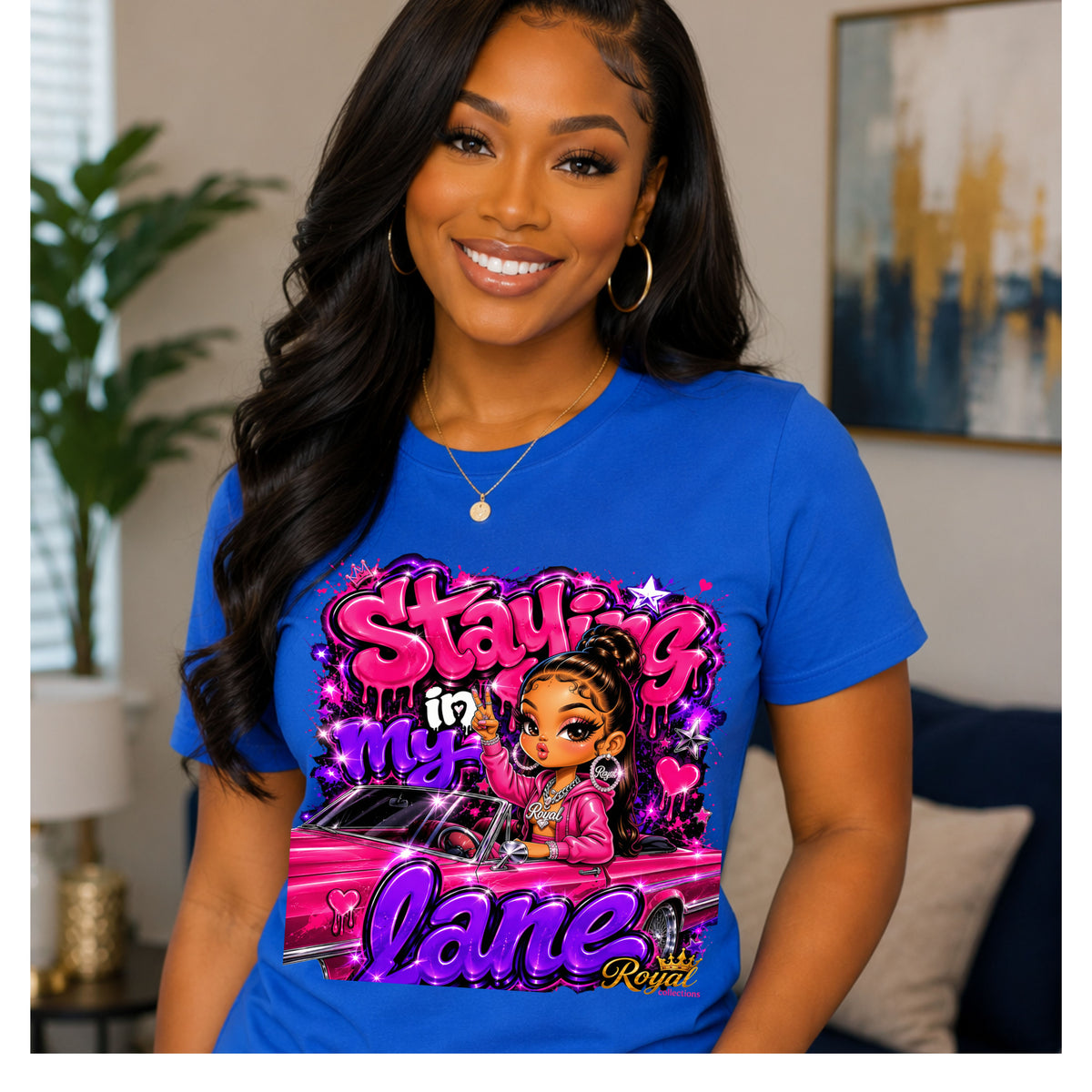A smiling woman wearing a bright blue graphic T-shirt with a colorful cartoon-style print that reads 'Staying in My Lane' and 'Kevina'. The photo is taken indoors with a blurred living room background.