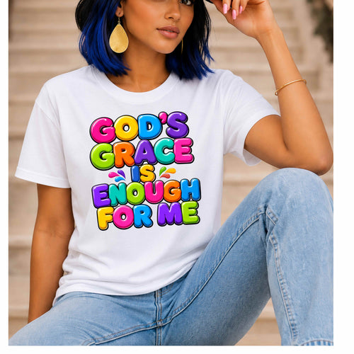 A woman wearing a white graphic T-shirt with colorful bubble text reading 'GOD'S GRACE IS ENOUGH FOR ME', paired with light blue jeans, seated against a softly blurred beige background.