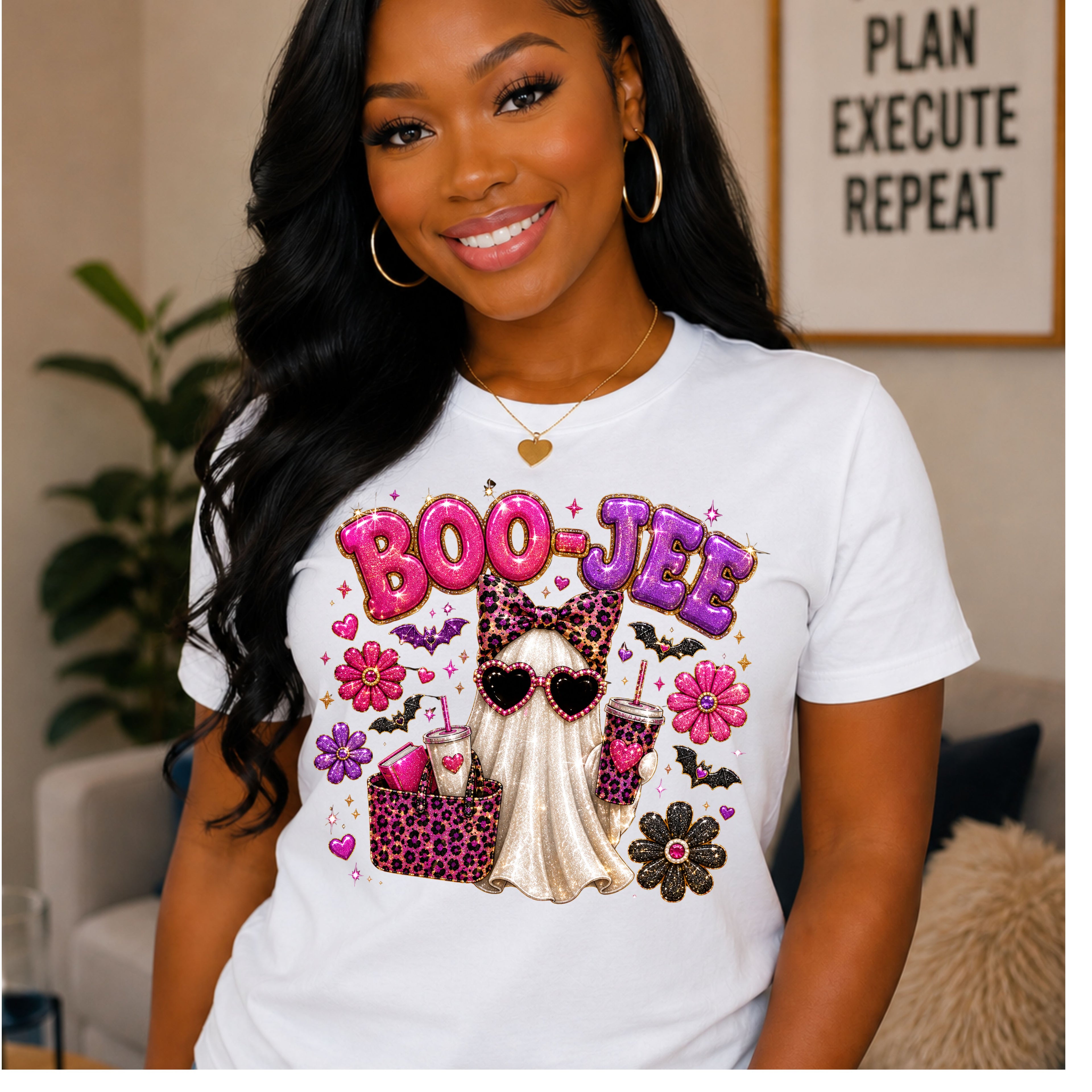 A woman wearing a white graphic t-shirt with a colorful Halloween-themed design featuring the text 'BOO-JEE' in pink and purple, a ghost in leopard print, flowers, and drinks. The background shows a cozy indoor setting with a framed sign that reads 'PLAN EXECUTE REPEAT'.