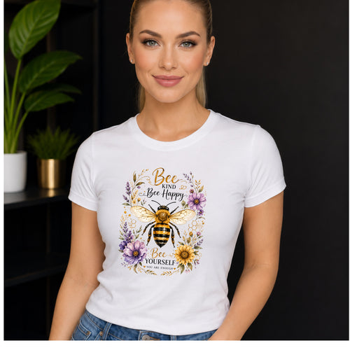 A woman wearing a white short-sleeve t-shirt with a colorful graphic of a bumblebee and flowers, set against a dark background with a blurred green plant. "Bee Kind, Bee Happy" t-shirt