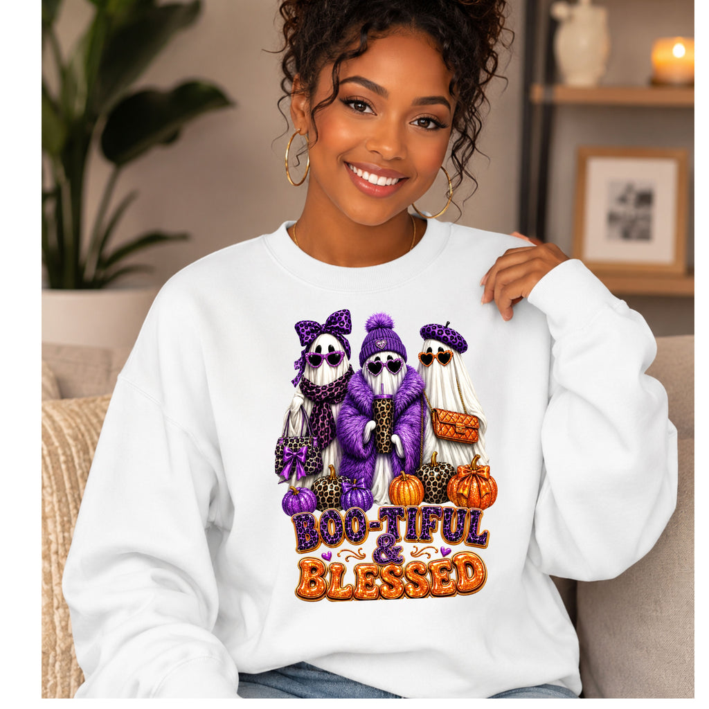 A woman wearing a white crewneck sweatshirt with a colorful graphic of three ghosts in purple and orange Halloween outfits, with the text 'BOO-TIFUL & BLESSED' below them, set in a cozy indoor environment.
