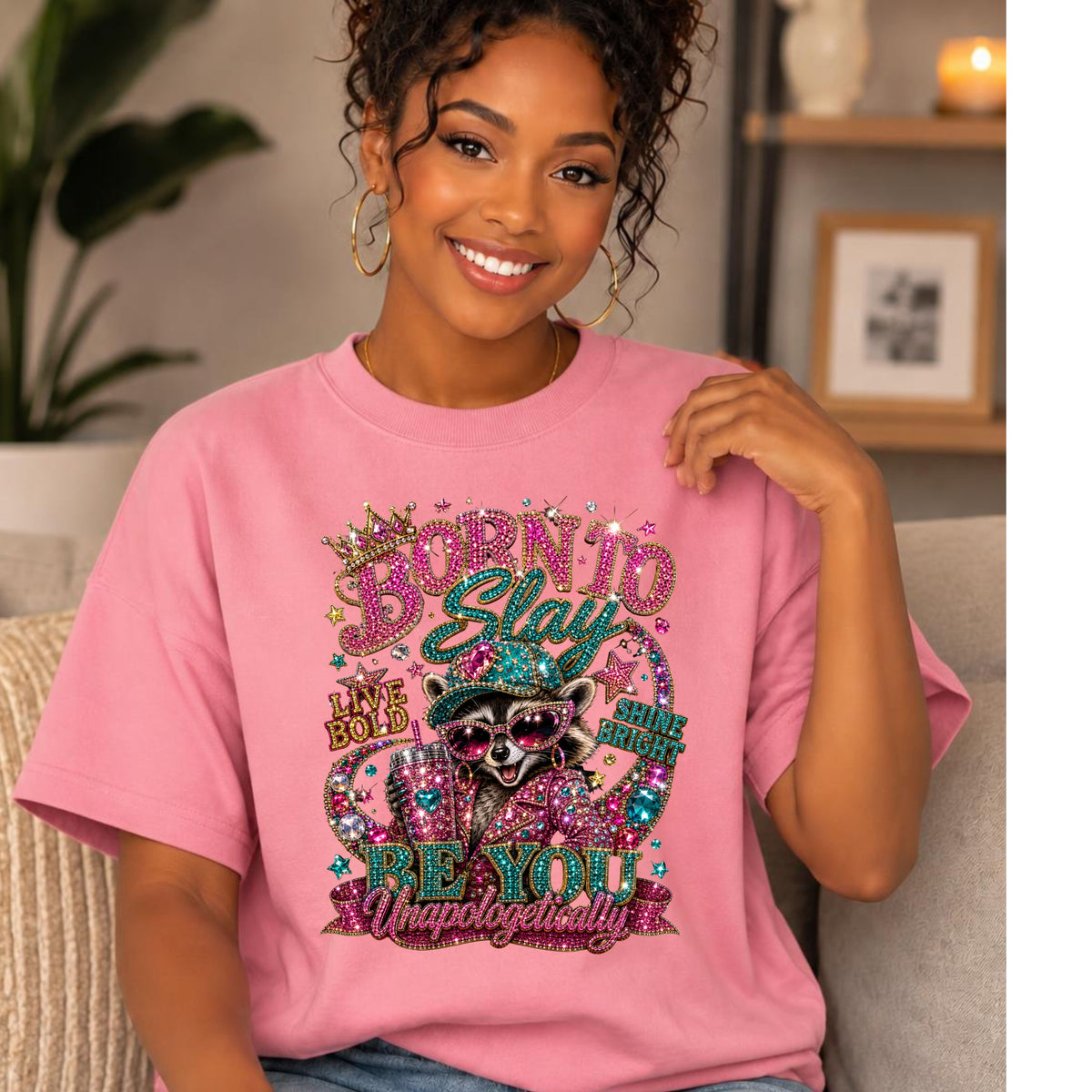 A woman wearing a pink graphic t-shirt with a rhinestone-style raccoon design and the text 'BORN TO STAY BE YOU Unhappily'.