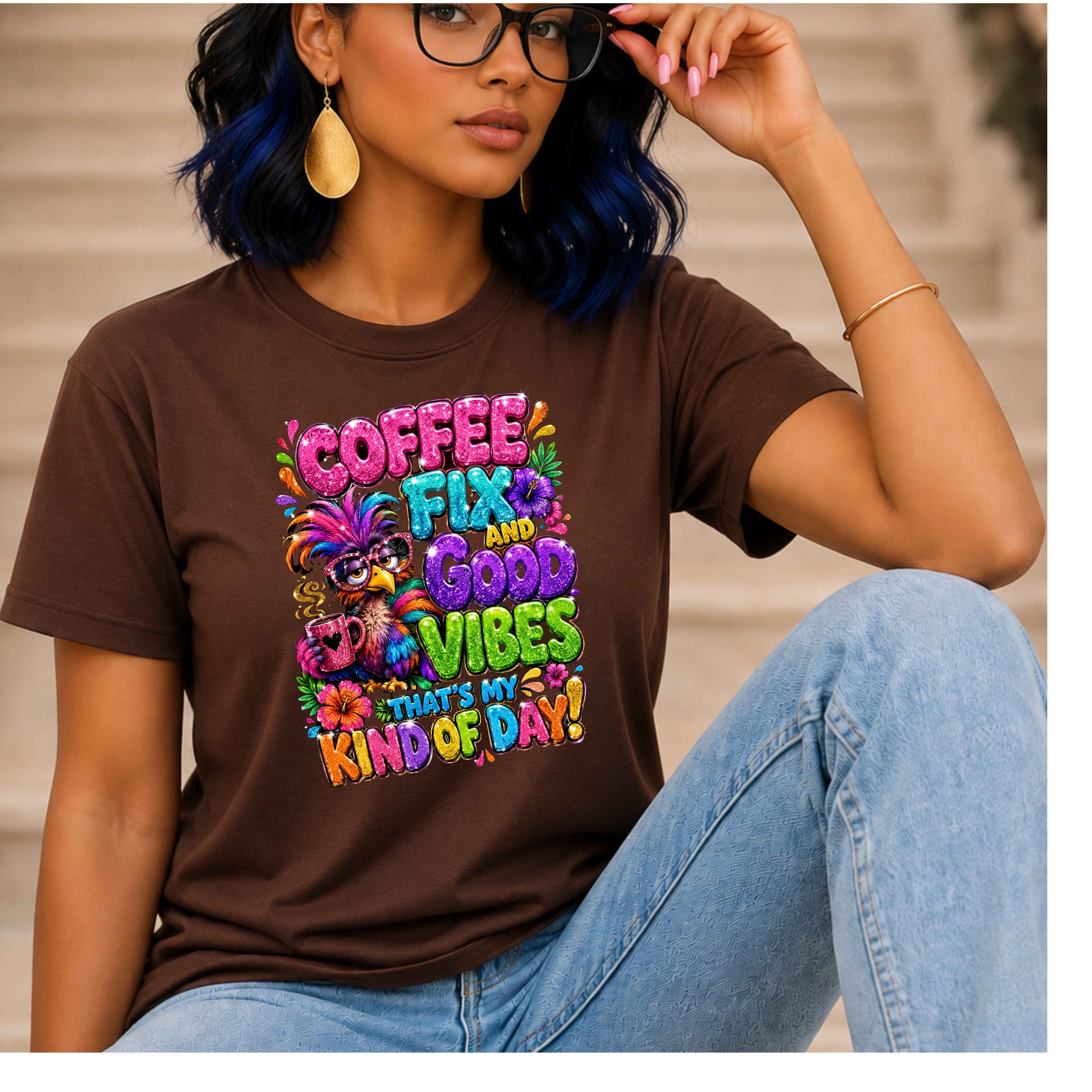 A woman wearing a brown graphic T-shirt with colorful text reading 'COFFEE FIX AND GOOD VIBES THAT'S MY KIND OF DAY!' and a cartoon character, paired with light blue jeans, seated in front of a blurred beige background.