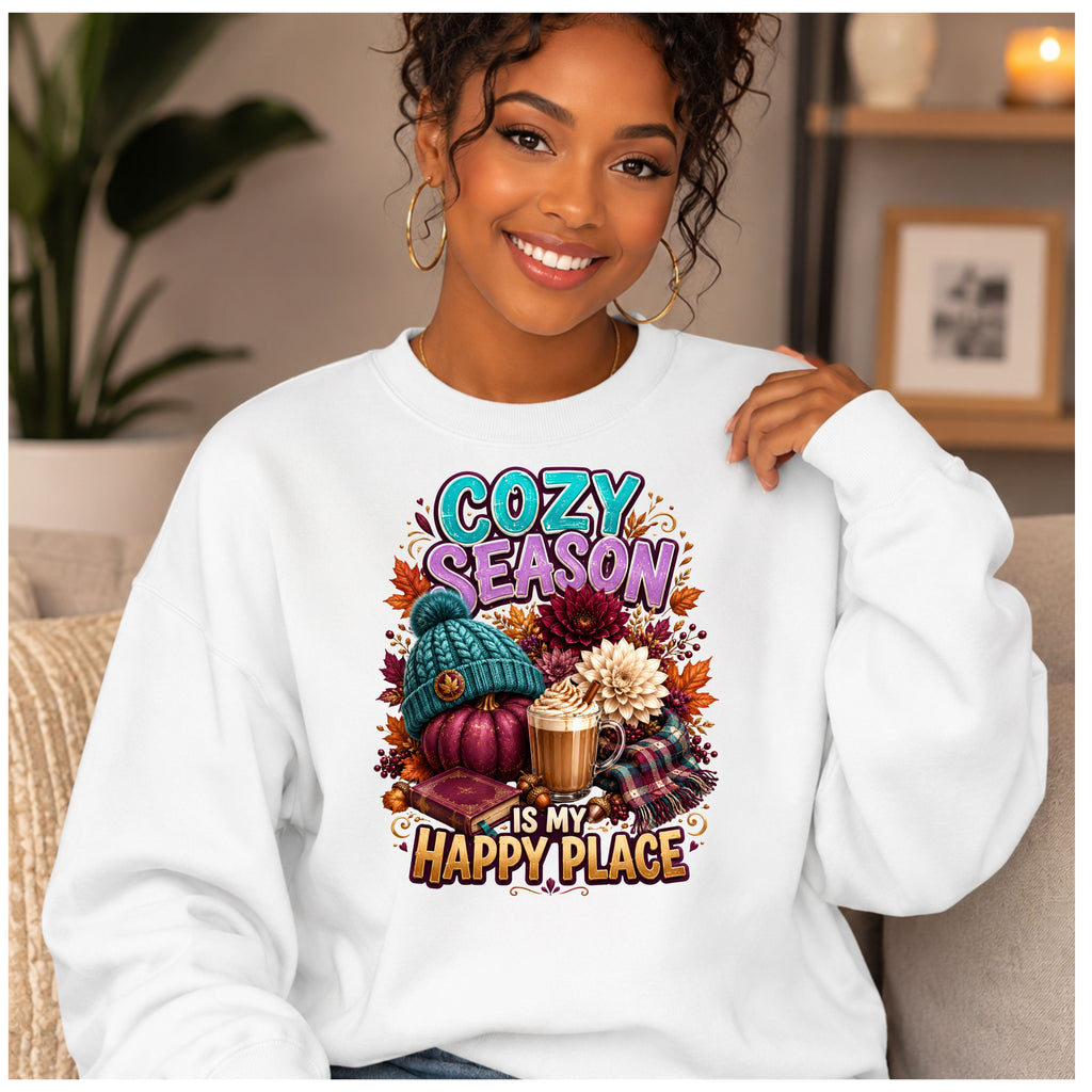 A smiling woman wearing a white crewneck sweatshirt with a colorful autumn-themed graphic that reads 'COZY SEASON IS MY HAPPY PLACE' above and below a design featuring a pumpkin, knit hat, coffee cup, flowers, and fall leaves.