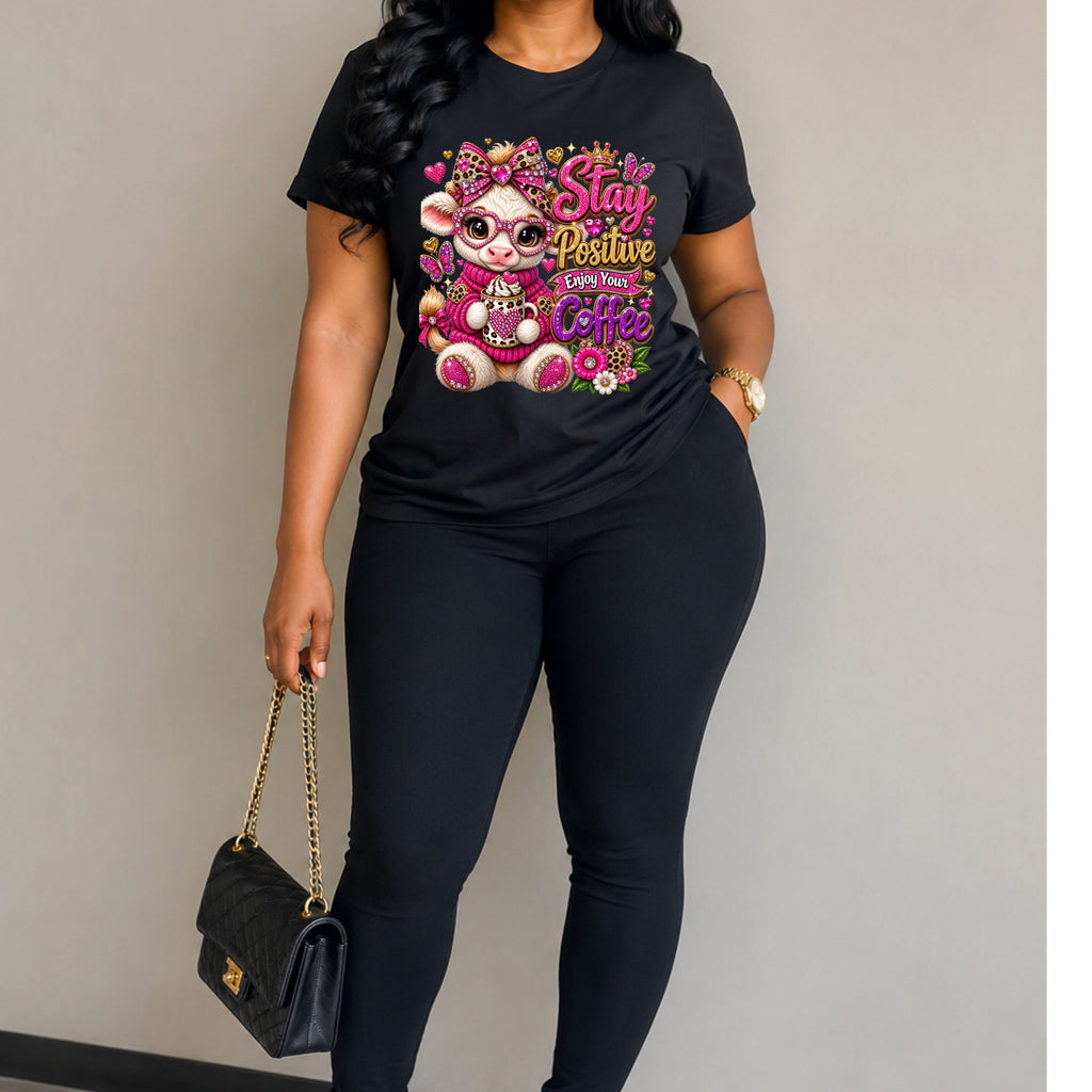 A woman wearing a black graphic t-shirt with a colorful cartoon bear and the text 'Stay Positive Love Your Coffee' paired with black leggings, holding a black quilted handbag with a gold chain strap against a plain gray wall.