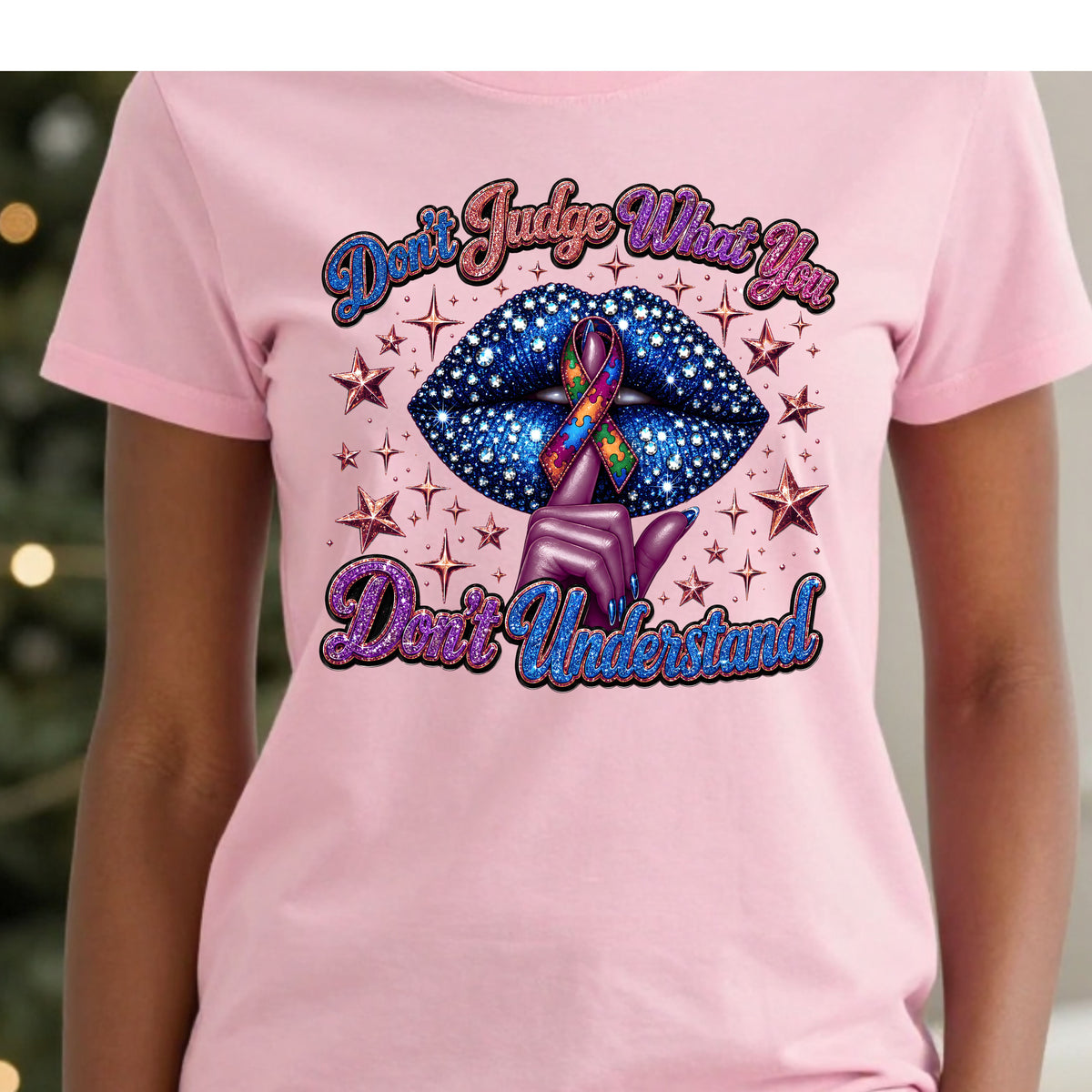 A person wearing a light pink short-sleeve T-shirt with a large graphic on the front. The graphic features the text 'Don't Judge What You Don't Understand' in blue and purple script around a pair of glittery blue lips with a multicolor awareness ribbon and purple hand gesture, set against a blurred holiday background.