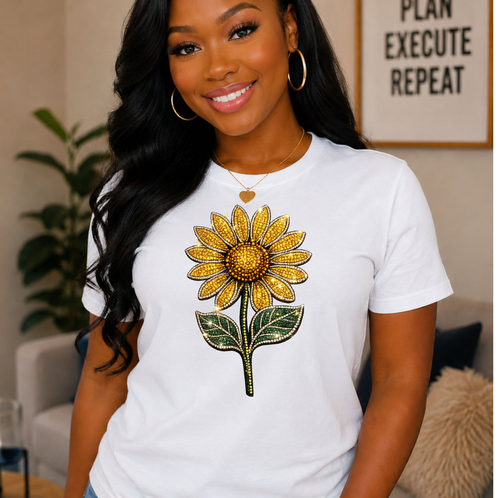 A woman wearing a white short-sleeve T-shirt with a large yellow sunflower graphic on the front, posed in a cozy indoor setting with a framed sign in the background.