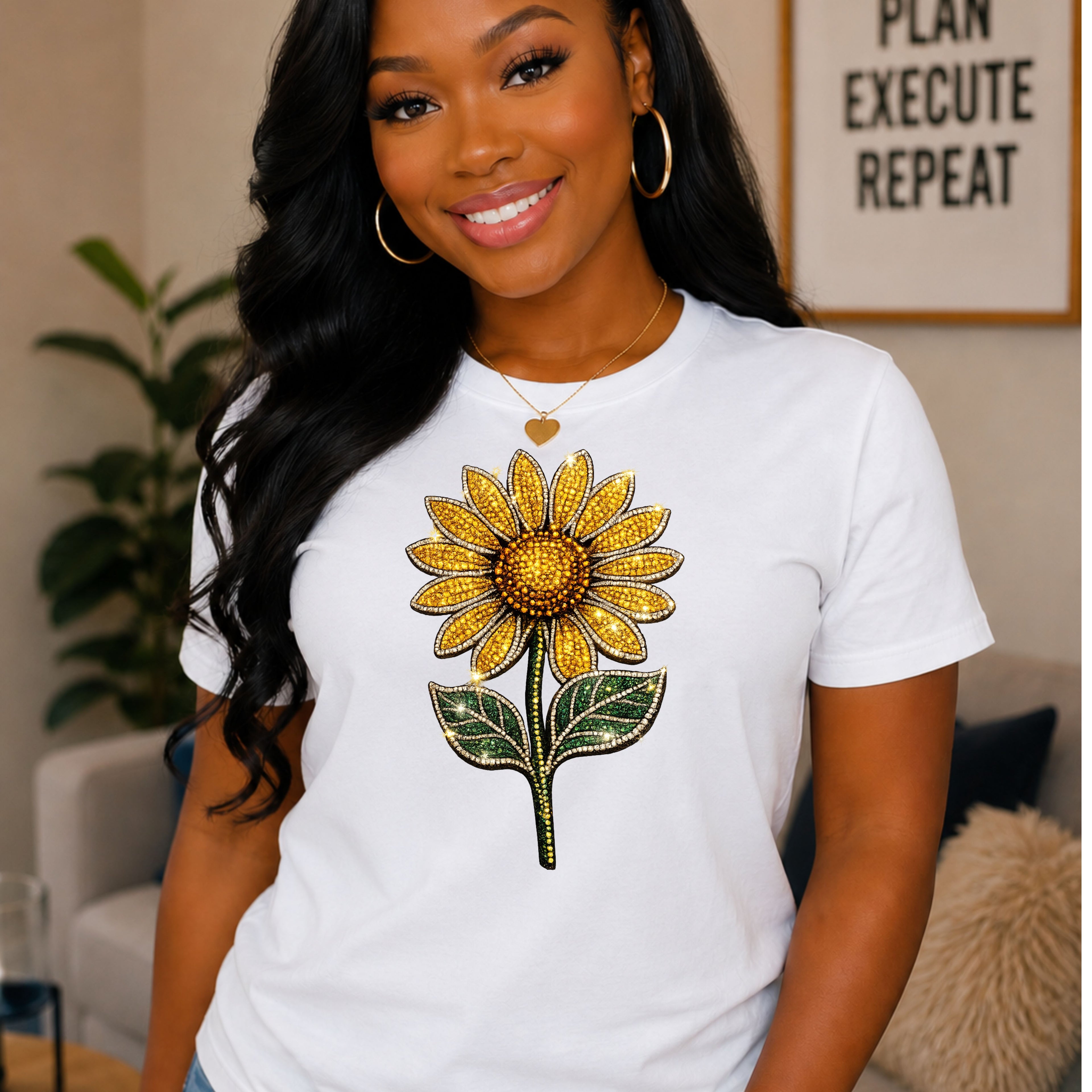 A woman wearing a white short-sleeve T-shirt with a large yellow sunflower graphic on the front, posed in a cozy indoor setting with a framed sign in the background.
