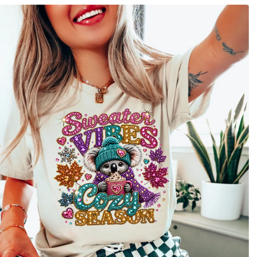 A woman wearing a cream-colored graphic t-shirt with a glittery, rhinestone-style design featuring a koala in a teal beanie and scarf, surrounded by the words 'Sweater VIBES Cozy SEASON'.