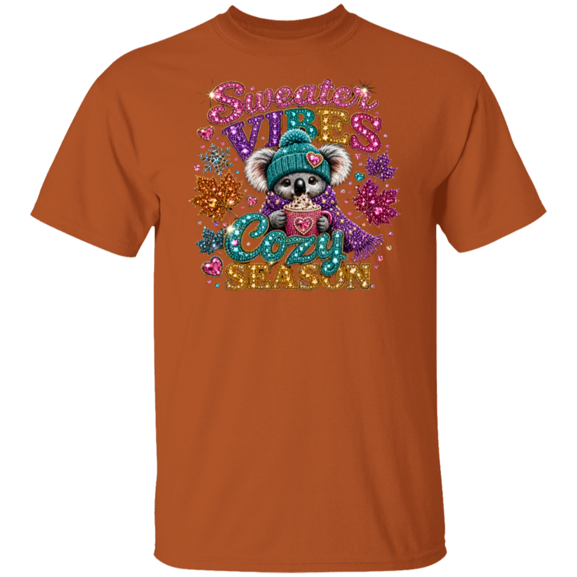 An orange crewneck t-shirt featuring a colorful, glittery graphic of a koala wearing a teal beanie and holding a heart-shaped mug, surrounded by the text 'Sweeter VIBES Cozy SEASON' and decorative snowflakes.