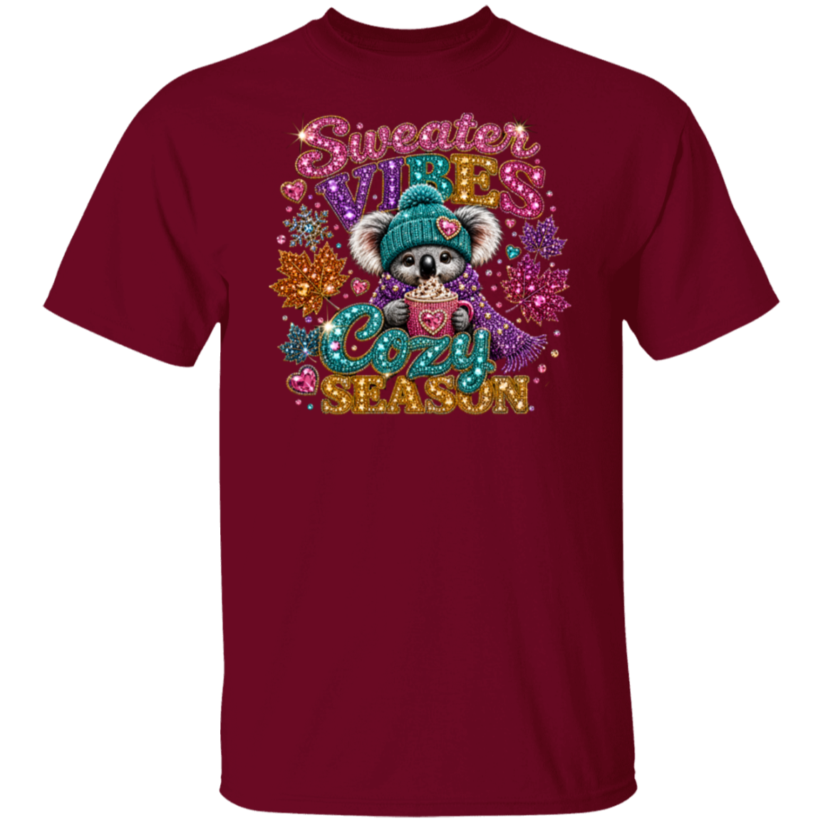 A garnet color short-sleeve crewneck t-shirt featuring a colorful graphic of a koala wearing a teal beanie and holding a mug, surrounded by glittery text that reads 'Sweater Vibes Cozy Season' and autumn leaves.