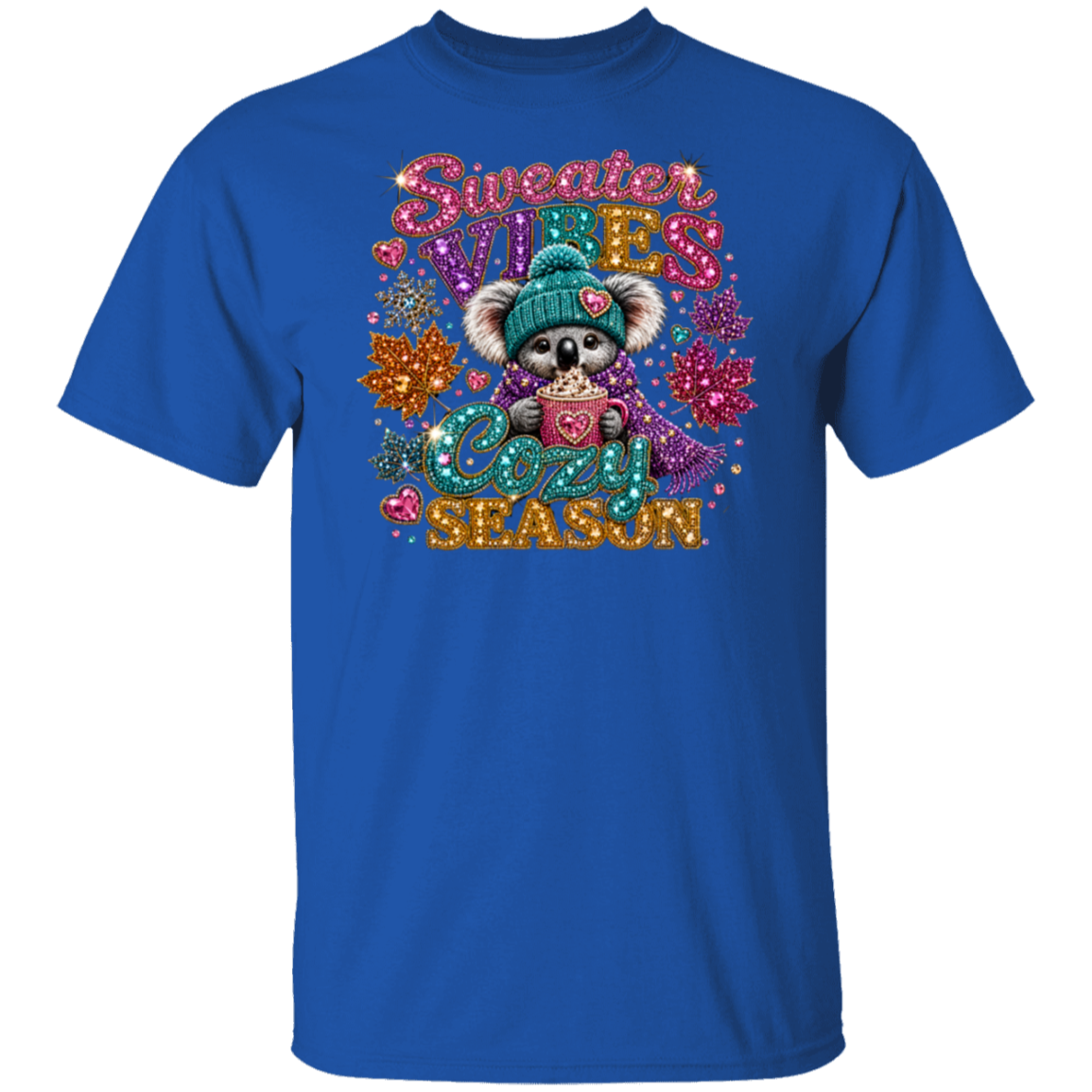 A royal blue short-sleeve crewneck t-shirt featuring a colorful graphic print of a cute koala wearing a teal knit hat and scarf, surrounded by glittery text that reads 'Sweater VILES SEASON' and winter-themed embellishments.