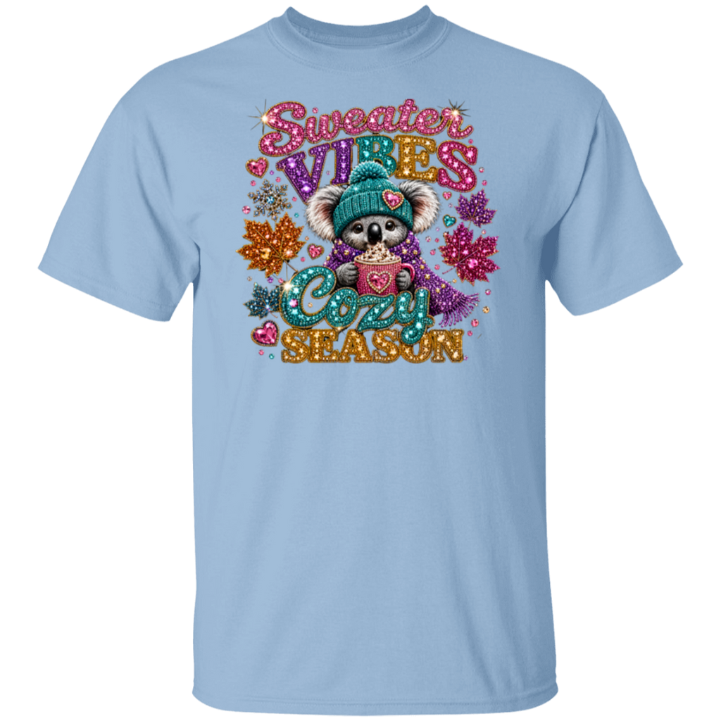 A light blue short-sleeve crewneck t-shirt featuring a colorful graphic of a cute cartoon koala wearing a teal knit hat and scarf, holding a mug with 'COZY' written inside. The graphic is surrounded by glittery text reading 'Sweater VIBES COZY SEASON' and autumn-themed decorations, shown against a plain white background.