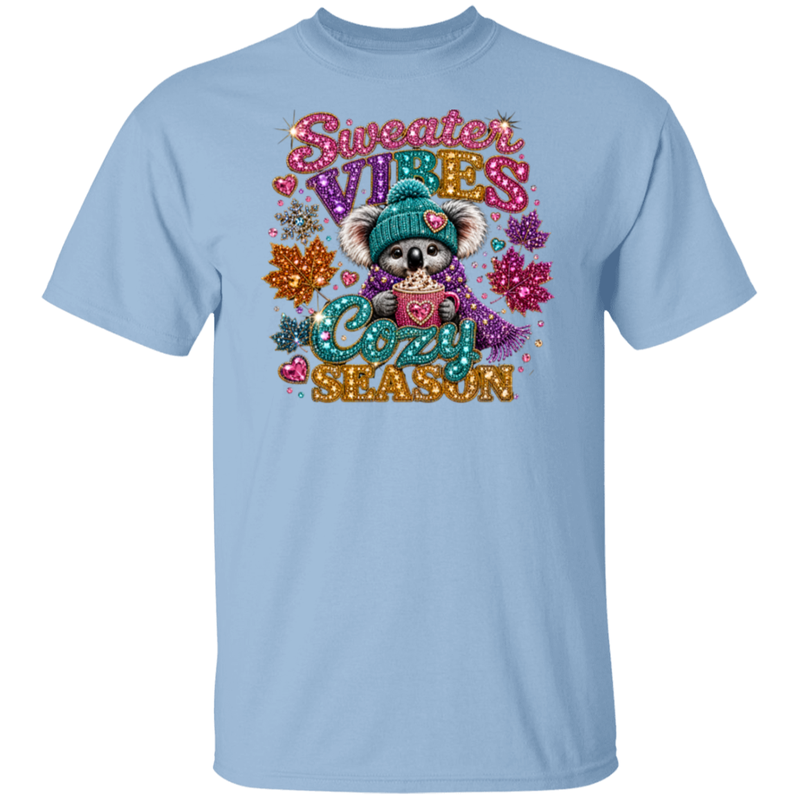 A light blue short-sleeve crewneck t-shirt featuring a colorful graphic of a cute cartoon koala wearing a teal knit hat and scarf, holding a mug with 'COZY' written inside. The graphic is surrounded by glittery text reading 'Sweater VIBES COZY SEASON' and autumn-themed decorations, shown against a plain white background.