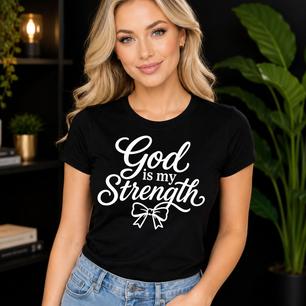 A blonde woman wearing a black color graphic t-shirt with the white text 'God is my Strength' and a bow illustration, paired with light blue jeans, set against a dark background with indoor plants.
