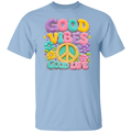 A light blue short-sleeve crewneck t-shirt featuring a colorful retro graphic on the chest with the text 'GOOD VIBES GOOD LIFE' in pastel bubble letters and a peace sign surrounded by flowers.