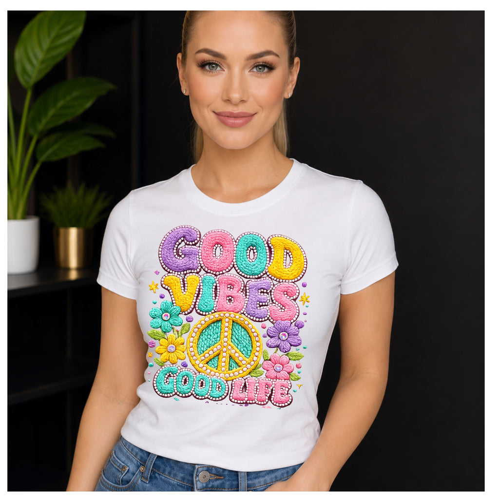 A woman wearing a white short-sleeve t-shirt with a colorful retro graphic that reads 'GOOD VIBES GOOD LIFE' with flowers, peace sign, and stars, posed against a dark indoor background with a plant.