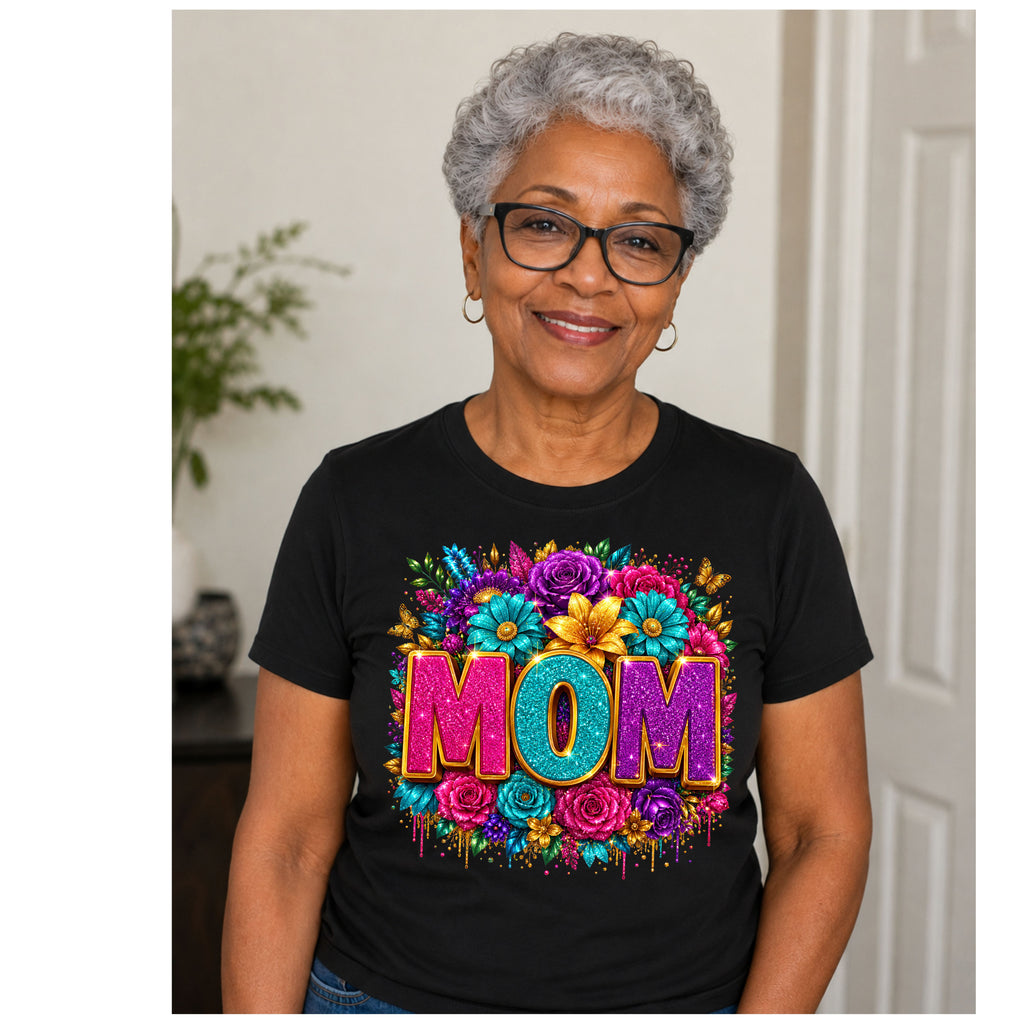 An older woman with short gray hair and glasses wearing a black graphic t-shirt with a colorful floral design and the word 'MOM' in glittery letters, standing indoors.