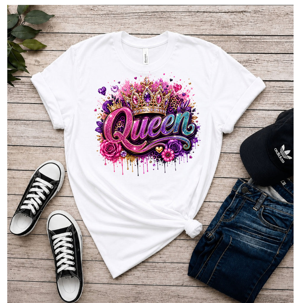 A white graphic t-shirt featuring a colorful 'Queen' text design with a crown, roses, and paint splatters, styled in a flat lay with black sneakers, blue jeans, and a black Adidas cap on a wooden background.