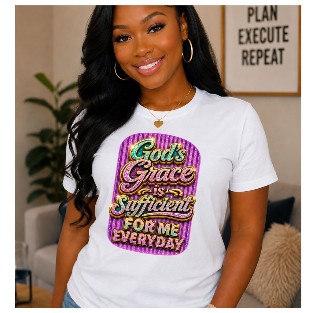 A woman wearing a white graphic t-shirt with a colorful text print that reads 'God's Grace is Sufficient FOR ME EVERYDAY' in a purple and pink striped design, posed in a living room setting.
