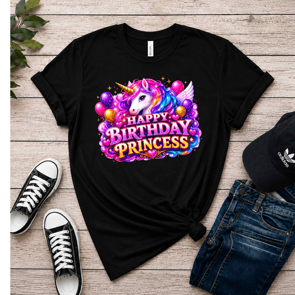 A black short-sleeve graphic T-shirt laid flat on a light wood surface, styled with black sneakers, blue jeans, and a black Adidas cap. The shirt features a large colorful unicorn and 'Happy Birthday Princess' graphic on the front.