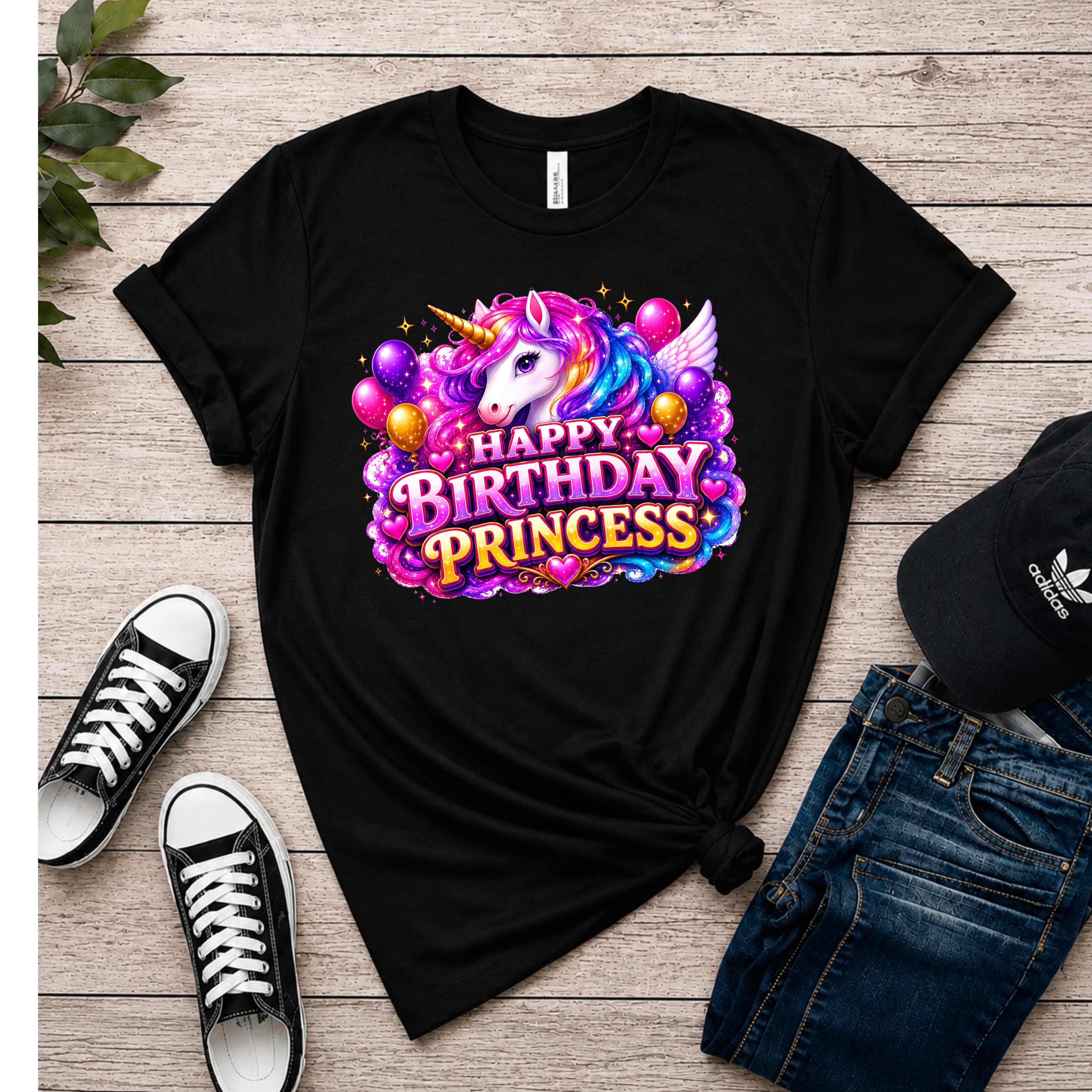 A black short-sleeve graphic T-shirt laid flat on a light wood surface, styled with black sneakers, blue jeans, and a black Adidas cap. The shirt features a large colorful unicorn and 'Happy Birthday Princess' graphic on the front.