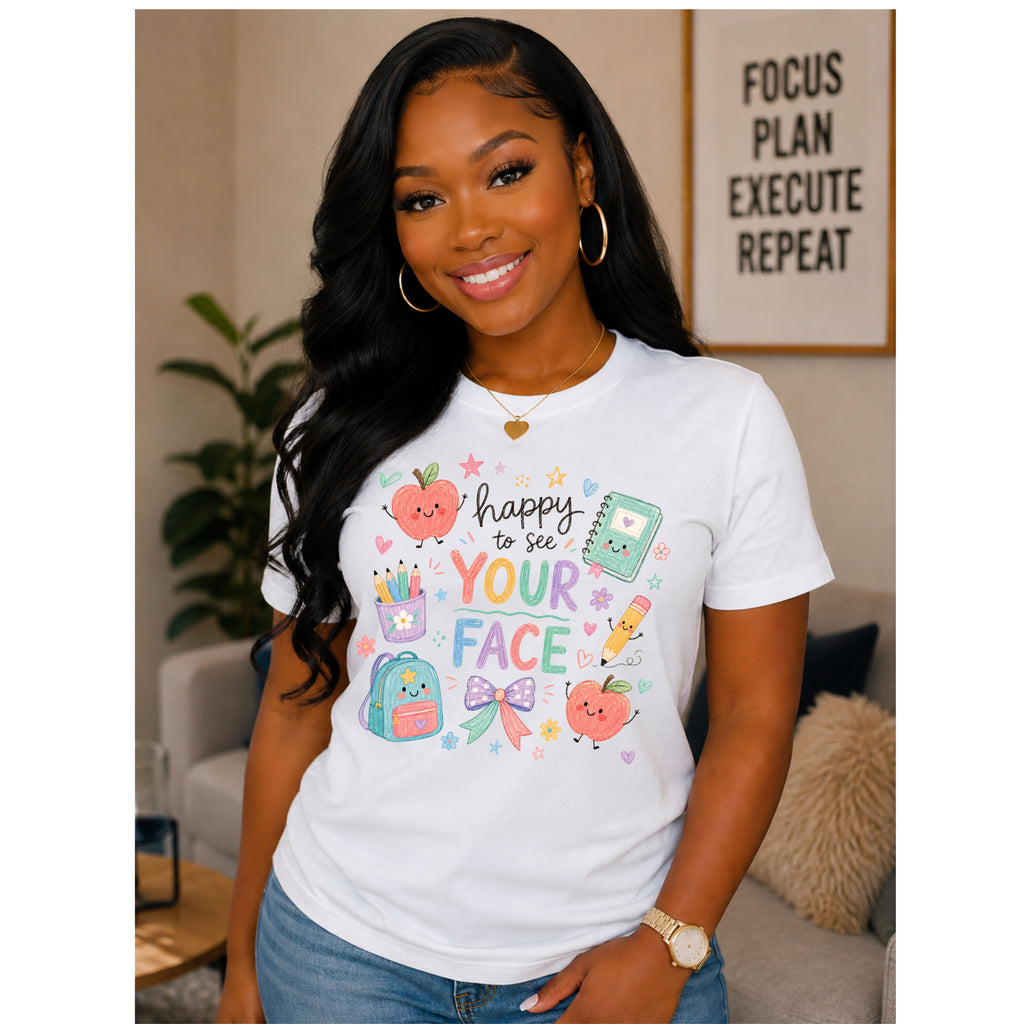 A woman wearing a white graphic t-shirt with a colorful school-themed design that reads 'happy to see YOUR FACE' with cute illustrations of a backpack, pencil cup, and apple. She is posing in a cozy living room setting.