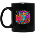 A glossy black ceramic mug featuring a vibrant, colorful graphic on the side. The graphic displays the word 'MOM' in glittery pink and purple letters, surrounded by a dense border of multicolored flowers including pink, blue, yellow, and purple, with a dripping paint effect at the bottom.