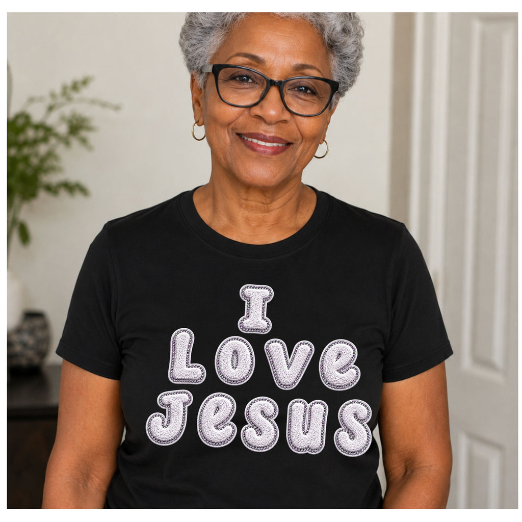 A smiling older woman with gray hair and glasses wearing a black short-sleeve T-shirt with the words 'I Love Jesus' in large white bubble letters outlined in gray, posed indoors against a light neutral wall.