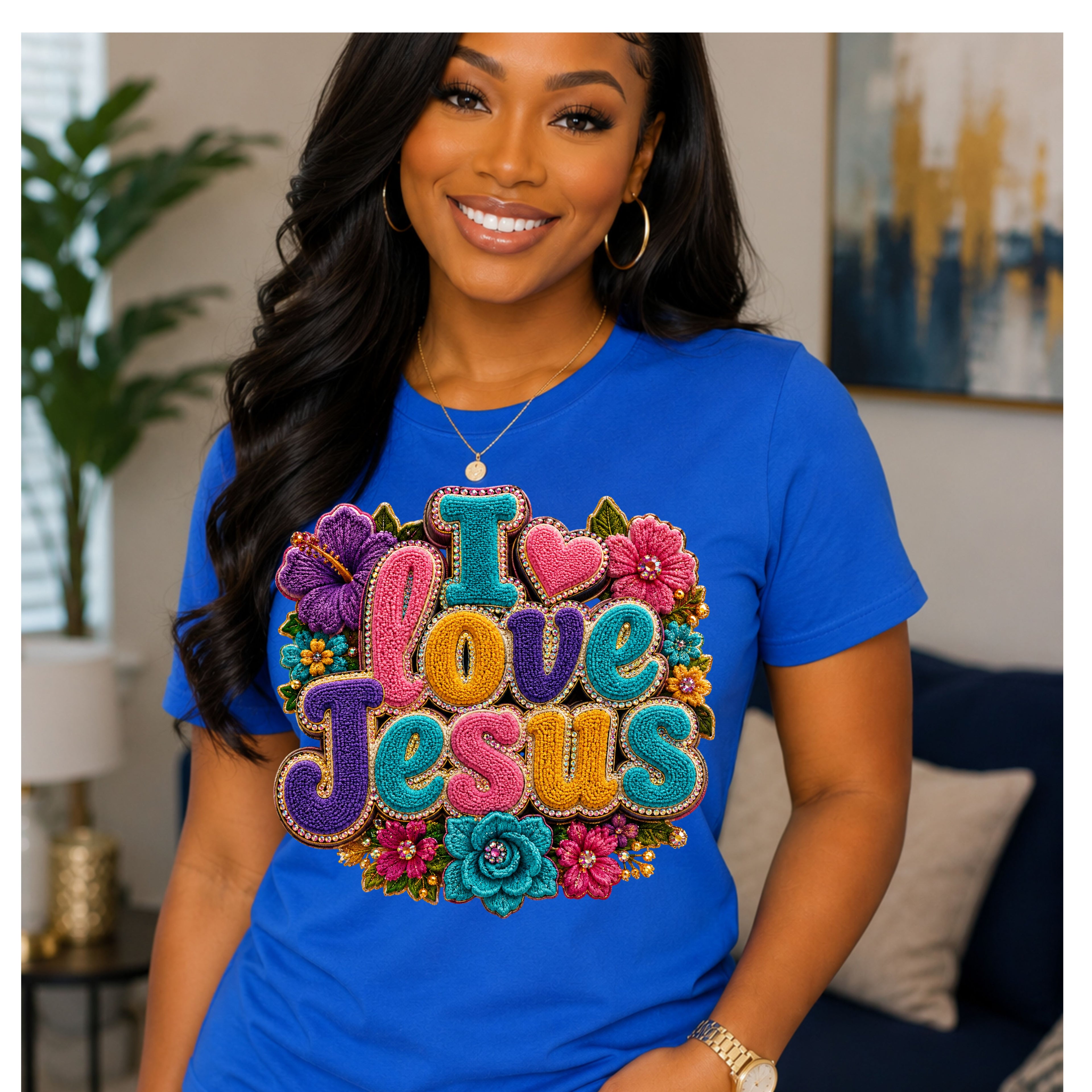 A smiling woman wearing a bright blue short-sleeve t-shirt with a large, colorful 3D-style graphic that reads 'I Love Jesus' surrounded by flowers. She is posing in a living room setting.