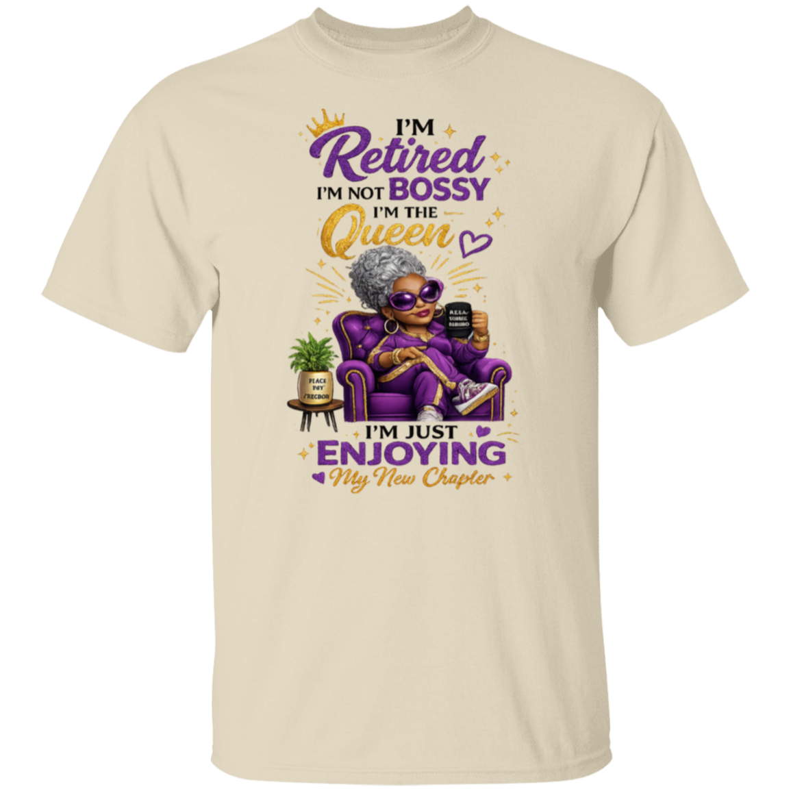 A natural color crewneck t-shirt featuring a colorful graphic of an elderly woman in a purple armchair with the text 'I'M Retired I'M NOT BOSSY I'M THE Queen I'm JUST ENJOYING My New Chapter'.