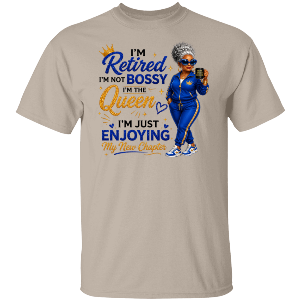 A sand color crewneck t-shirt featuring a colorful graphic print on the front. The graphic displays a cartoon illustration of a woman in a blue tracksuit holding a phone, next to the text 'I'm Retired I'm Not Bossy I'm The Queen I'm Just Enjoying My New Chapter' in blue and gold lettering.