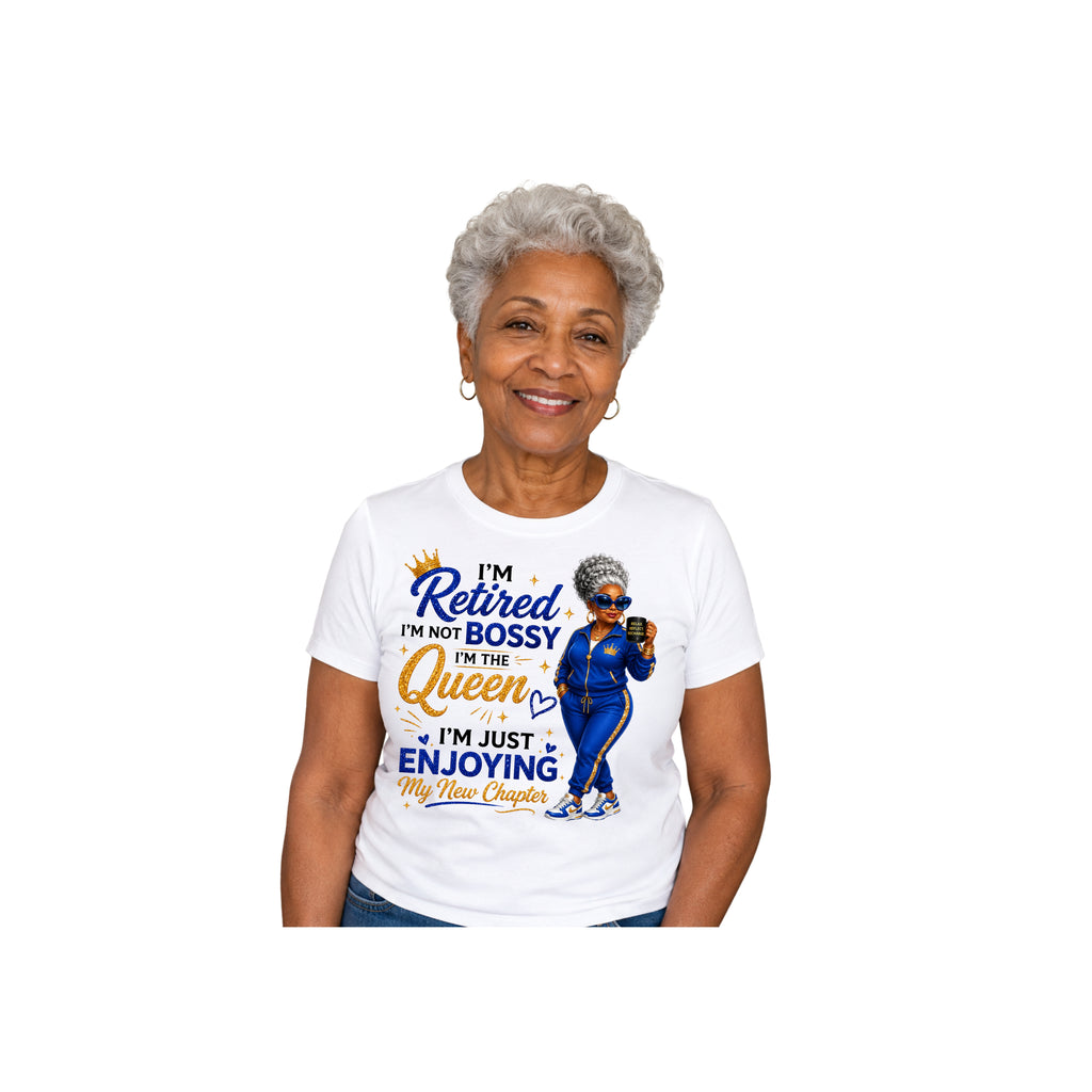An older woman with short gray hair is shown from the waist up wearing a white graphic T-shirt. The shirt features a blue and orange print with the text 'I'm Retired I'm Not Bossy In The Queen I'm Just Enjoying My New Chapter' and an illustration of a woman in a blue outfit.