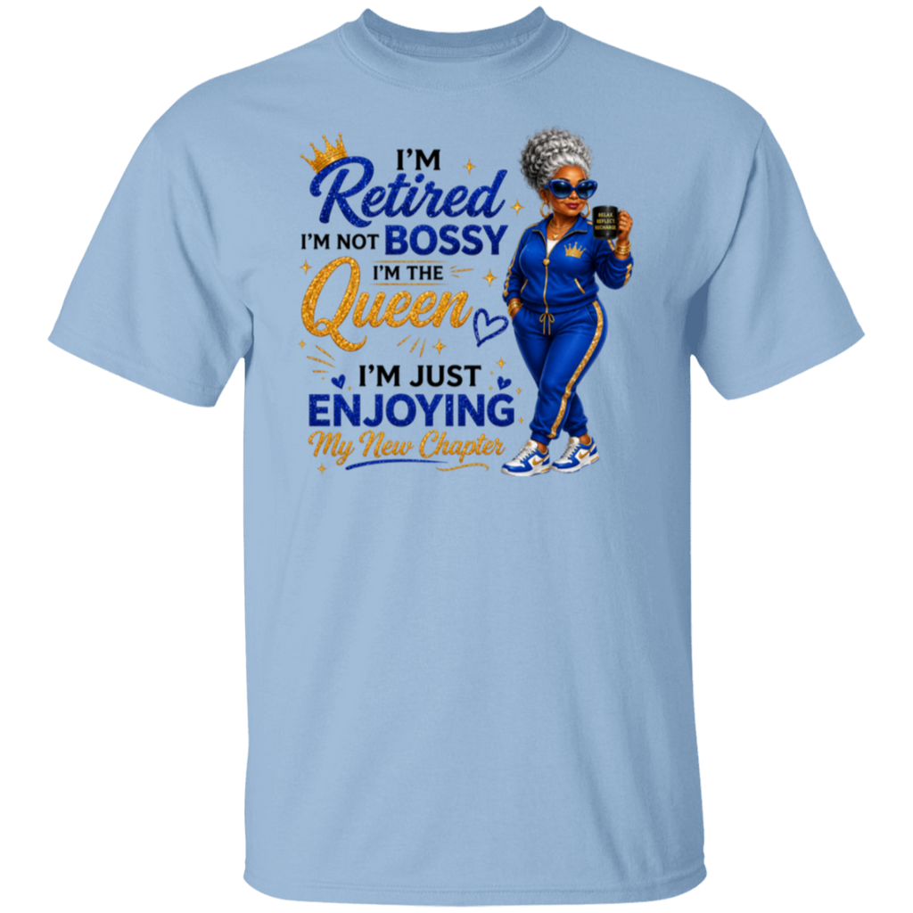 A light blue crewneck t-shirt featuring a colorful graphic print on the front. The graphic displays a cartoon illustration of a woman in a blue tracksuit holding a phone, next to the text 'I'm Retired I'm Not Bossy I'm The Queen I'm Just Enjoying My New Chapter' in blue and gold lettering.