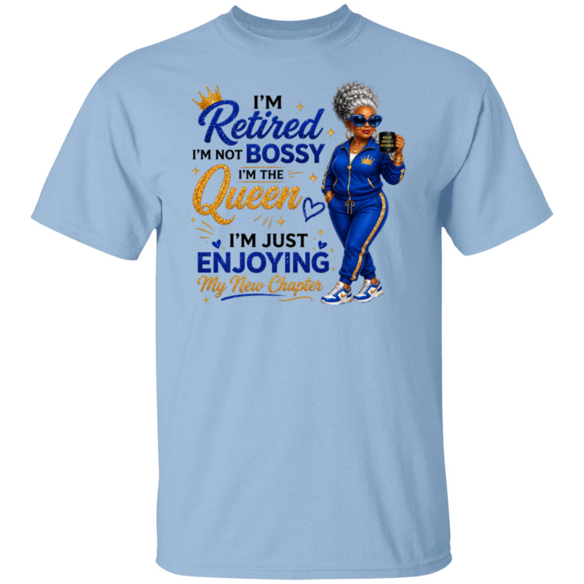A light blue crewneck t-shirt featuring a colorful graphic print on the front. The graphic displays a cartoon illustration of a woman in a blue tracksuit holding a phone, next to the text 'I'm Retired I'm Not Bossy I'm The Queen I'm Just Enjoying My New Chapter' in blue and gold lettering.