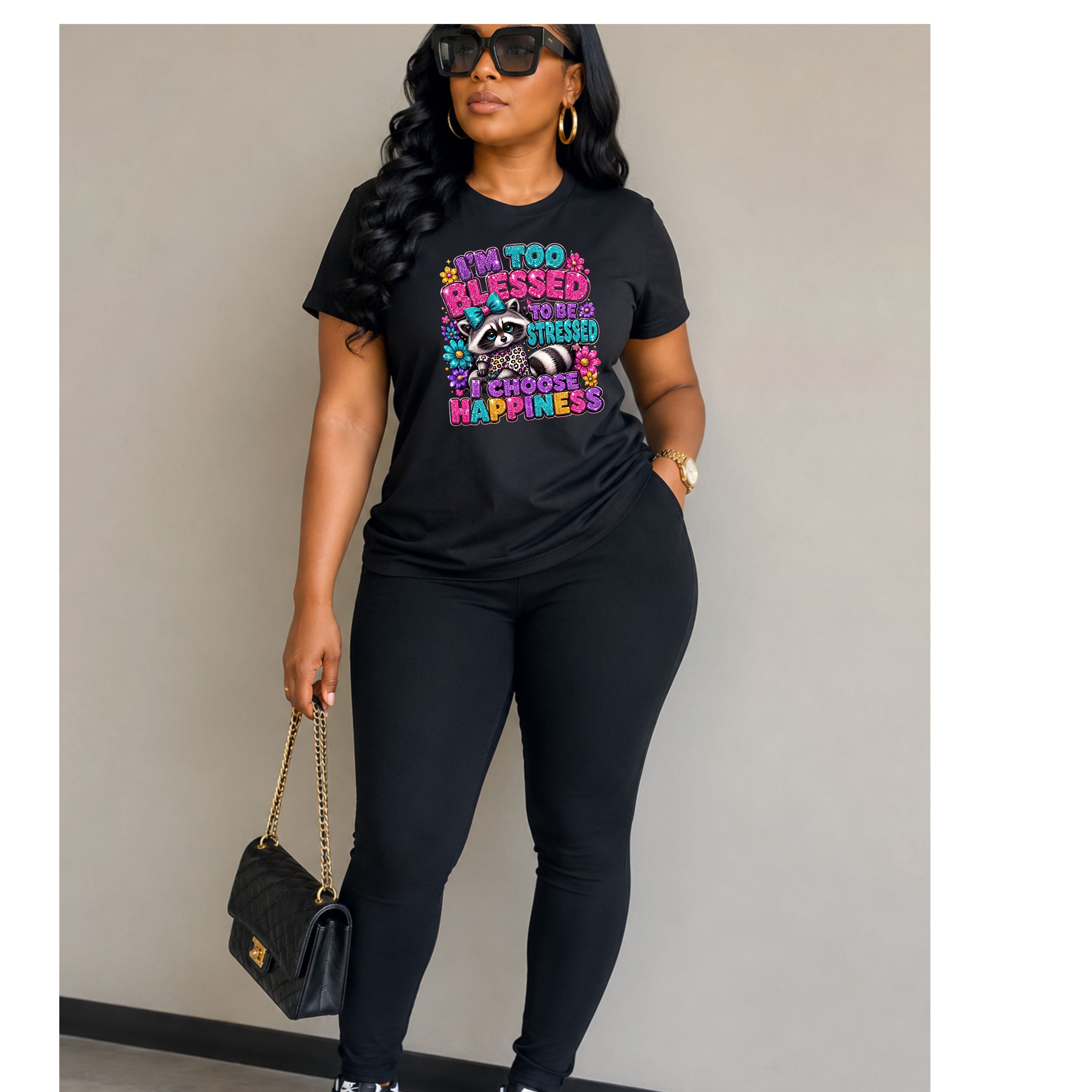 A woman modeling a black graphic t-shirt with a colorful raccoon design and the text 'I'M TOO BLESSED TO BE SAD. I'M TOO HAPPY TO BE SAD.