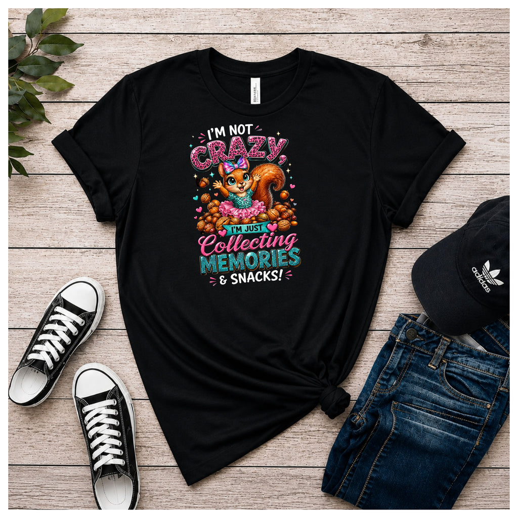 A black graphic t-shirt laid flat on a light wood background, styled with black sneakers, blue jeans, and a black Adidas cap. The t-shirt features a colorful graphic of a squirrel and the text 'I'M NOT CRAZY I'M JUST Collecting MEMORIES & SNACKS!'.