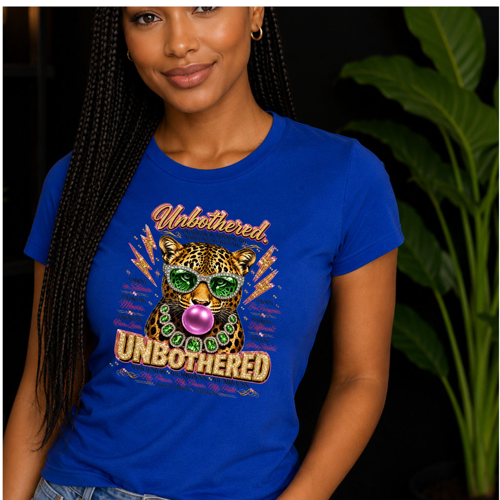A lady wearing a royal blue short-sleeve crewneck t-shirt featuring a colorful graphic print of a leopard wearing green sunglasses and a green beaded necklace, blowing a pink bubblegum bubble. The graphic includes the words 'Unbothered' and 'Unbothered' in stylized fonts, surrounded by motivational text while the lady is standing.