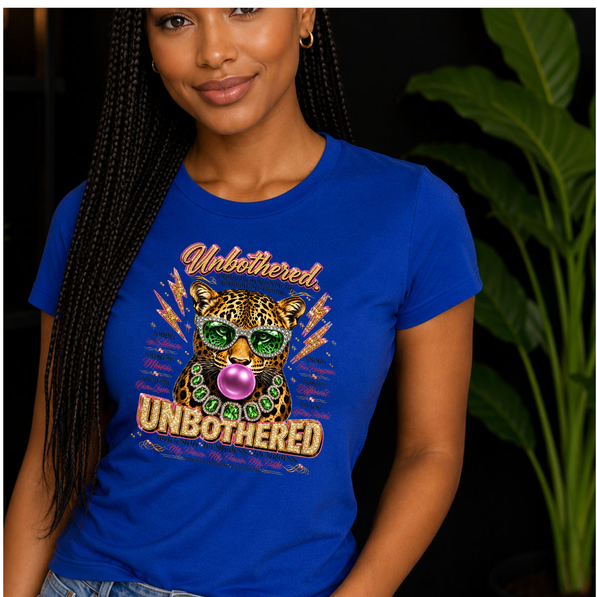 A lady wearing a royal blue short-sleeve crewneck t-shirt featuring a colorful graphic print of a leopard wearing green sunglasses and a green beaded necklace, blowing a pink bubblegum bubble. The graphic includes the words 'Unbothered' and 'Unbothered' in stylized fonts, surrounded by motivational text while the lady is standing.