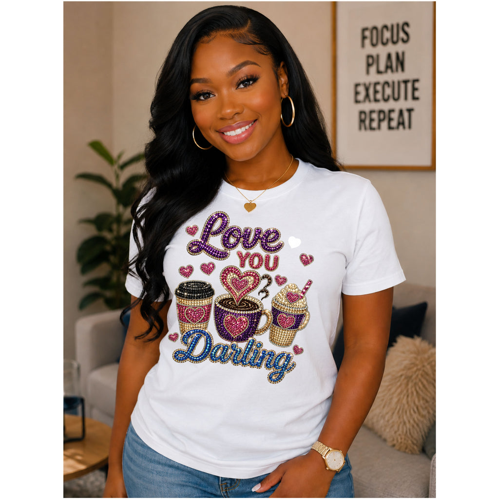 A woman wearing a white graphic t-shirt with 'Love You Darling' written in rhinestone-style lettering alongside coffee cup and cupcake illustrations, posed in a living room setting.