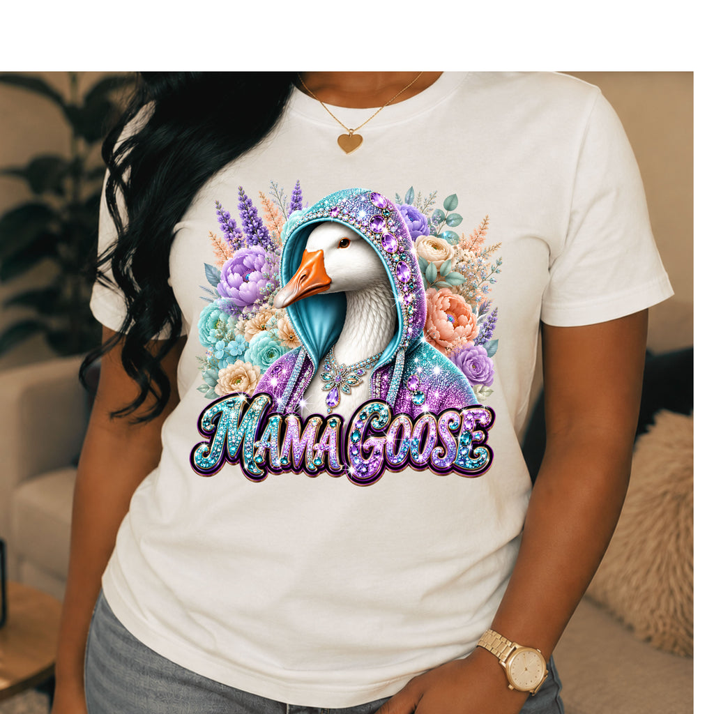 A woman wearing a white graphic t-shirt featuring a colorful illustration of a mother goose in a glittery purple hoodie surrounded by flowers, with the text 'MAMA GOOSE' below. The photo is taken in a cozy indoor setting.