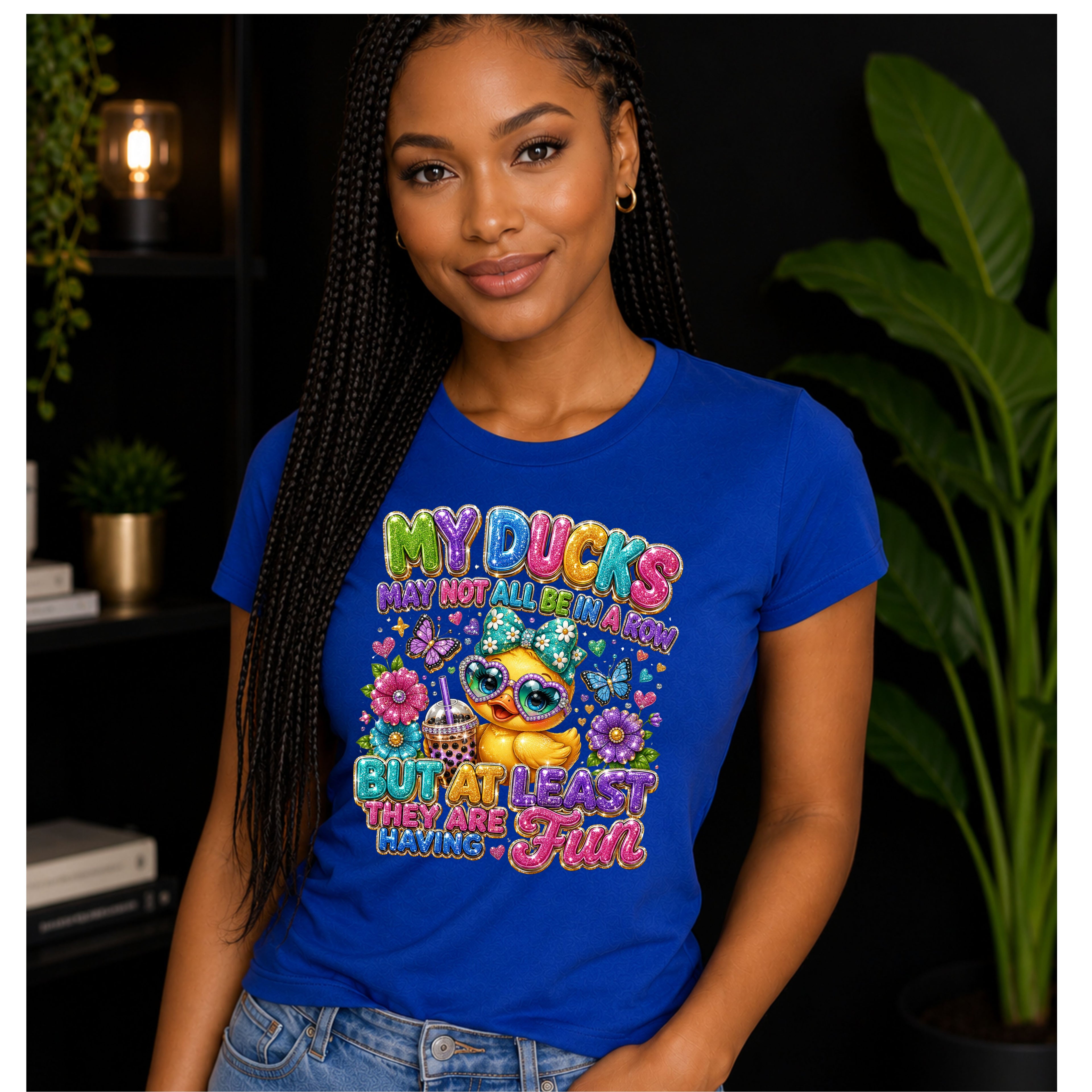 A woman wearing a blue graphic t-shirt with a colorful cartoon duck design and the text 'MY DUCKS MAY NOT ALL BE REAL BUT AT LEAST THEY ARE HAVING A Fun'. She is posing in front of a dark background with indoor plants.