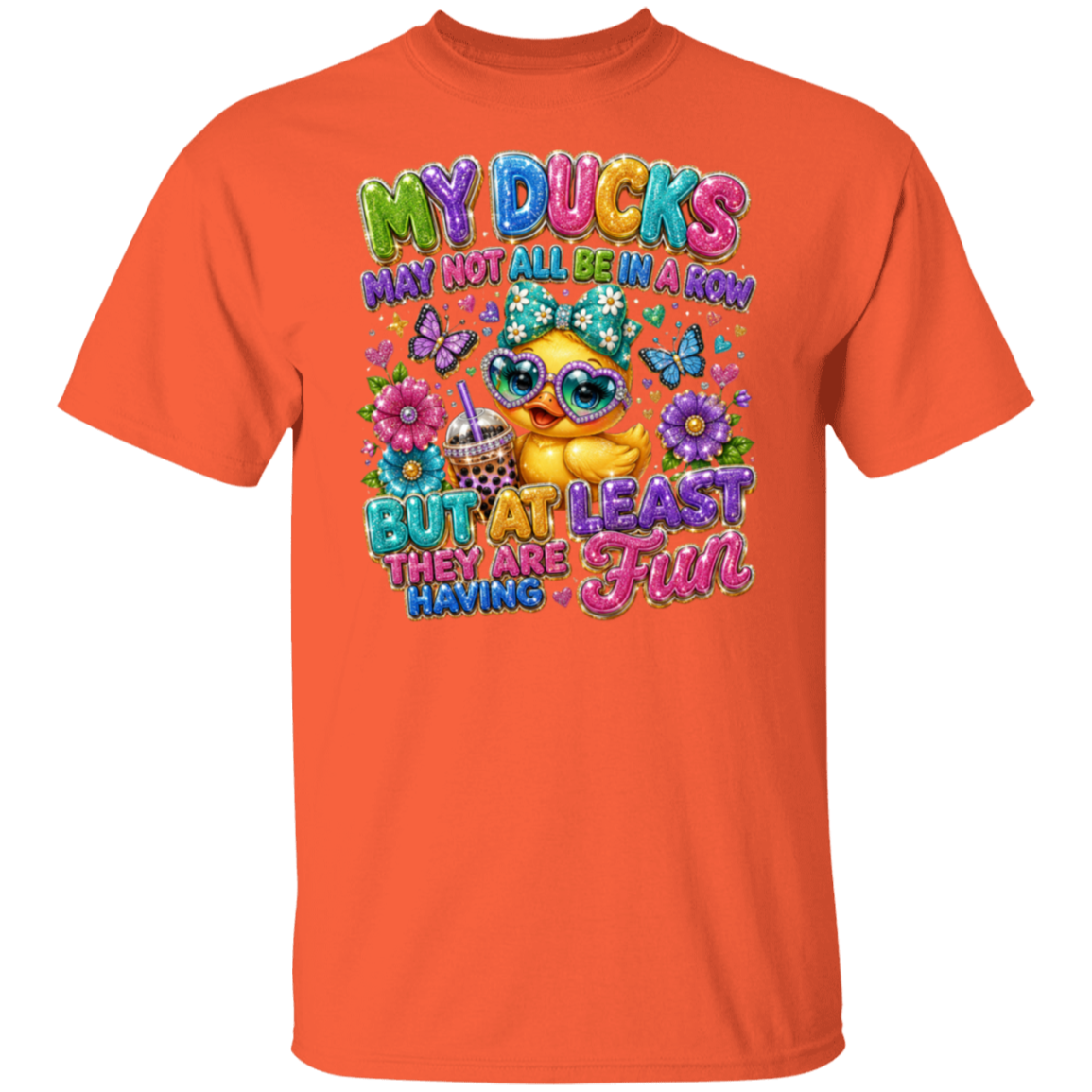An orange short-sleeve crewneck t-shirt featuring a colorful graphic print on the front. The graphic shows a yellow cartoon duck wearing sunglasses and a floral headband, surrounded by flowers, butterflies, and the text 'MY DUCKS MAY NOT ALL BE IN A RON BUT AT LEAST THEY ARE HAVING Fun'.