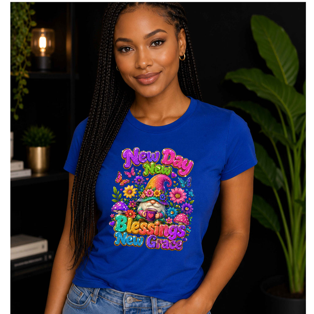 A woman wearing a royal blue graphic t-shirt with a colorful gnome and floral design that reads 'New Day New Grace' and 'Blessings New Grace', posed in front of a dark background with plants.