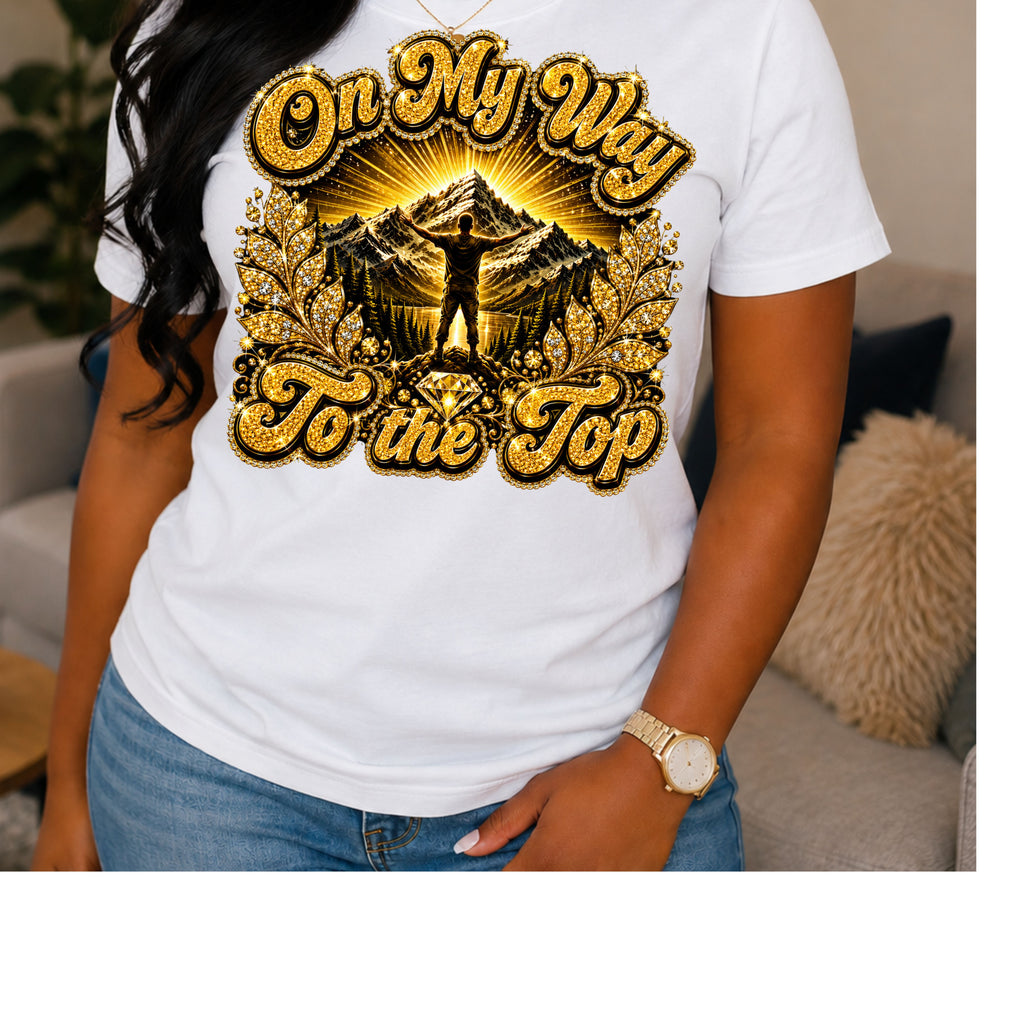A woman wearing a white short-sleeve graphic T-shirt with a large gold and black mountain design and the text 'On My Way To the Top' on the front, paired with blue jeans in a living room setting.