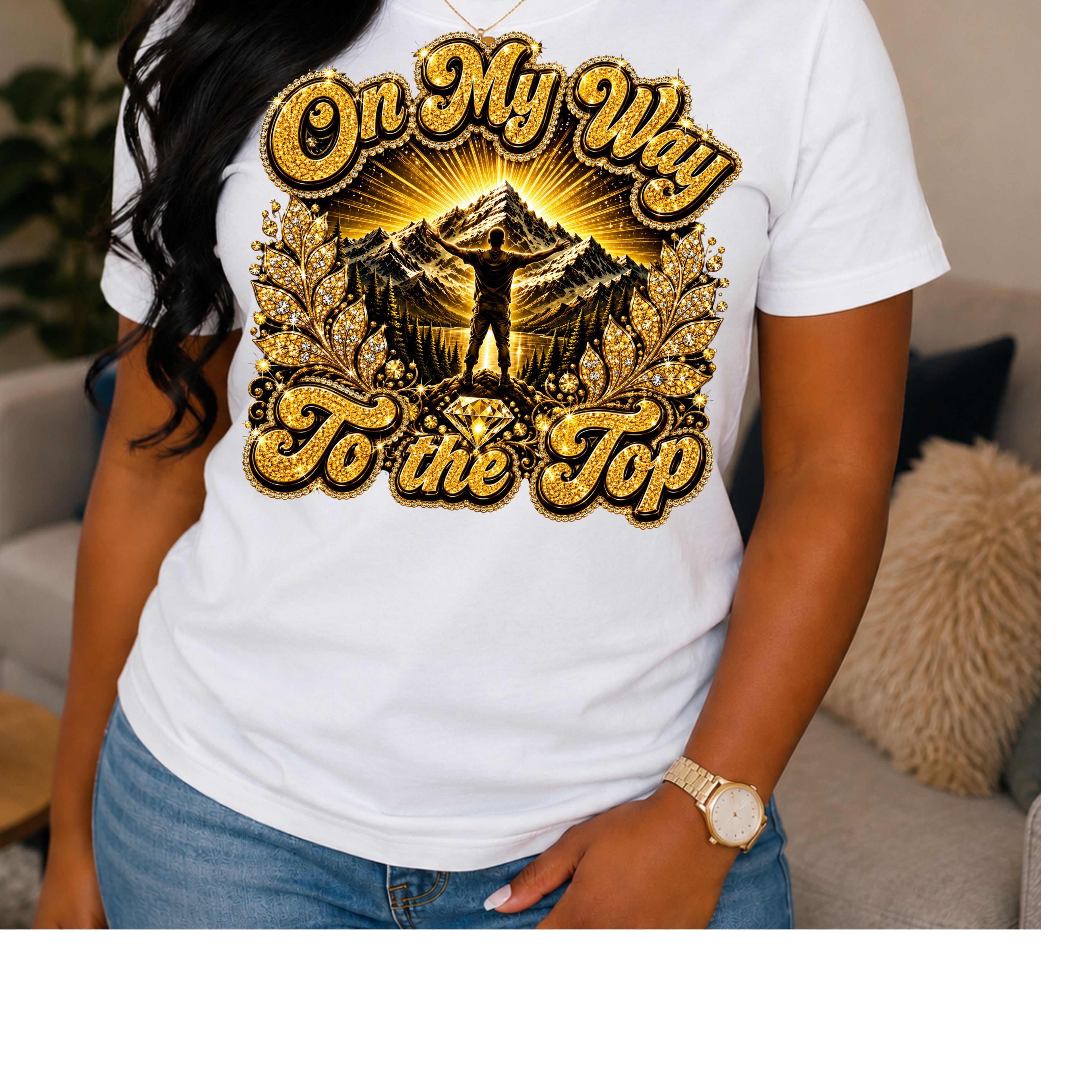 A woman wearing a white short-sleeve graphic T-shirt with a large gold and black mountain design and the text 'On My Way To the Top' on the front, paired with blue jeans in a living room setting.