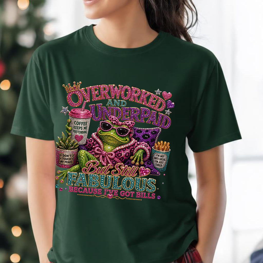 A woman wearing a dark green forest graphic t-shirt with a colorful, glittery design featuring a frog, Mardi Gras masks, and text that reads 'Overworked and Underpaid But Still Fabulous Because We Got Bills'.