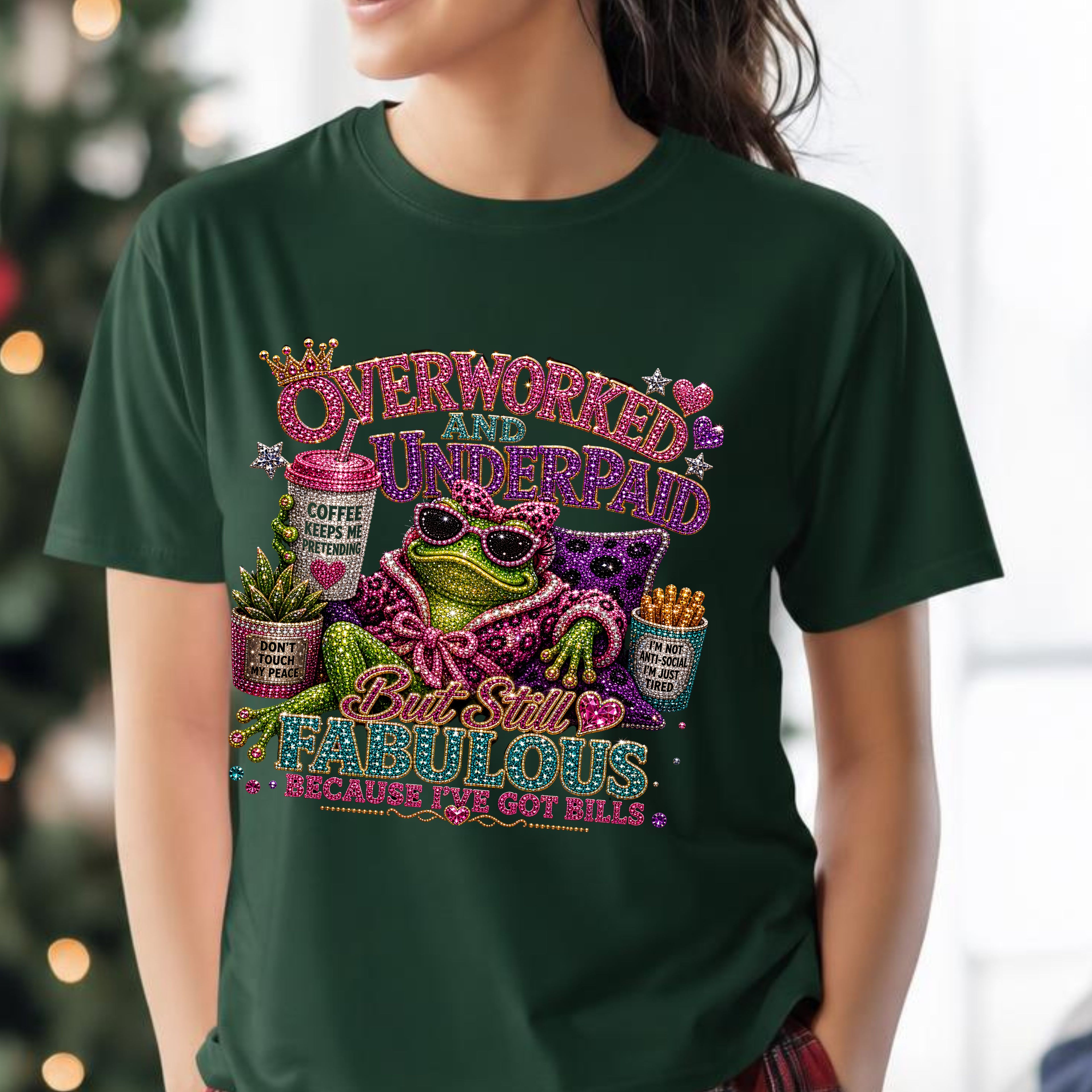 A woman wearing a dark green forest graphic t-shirt with a colorful, glittery design featuring a frog, Mardi Gras masks, and text that reads 'Overworked and Underpaid But Still Fabulous Because We Got Bills'.