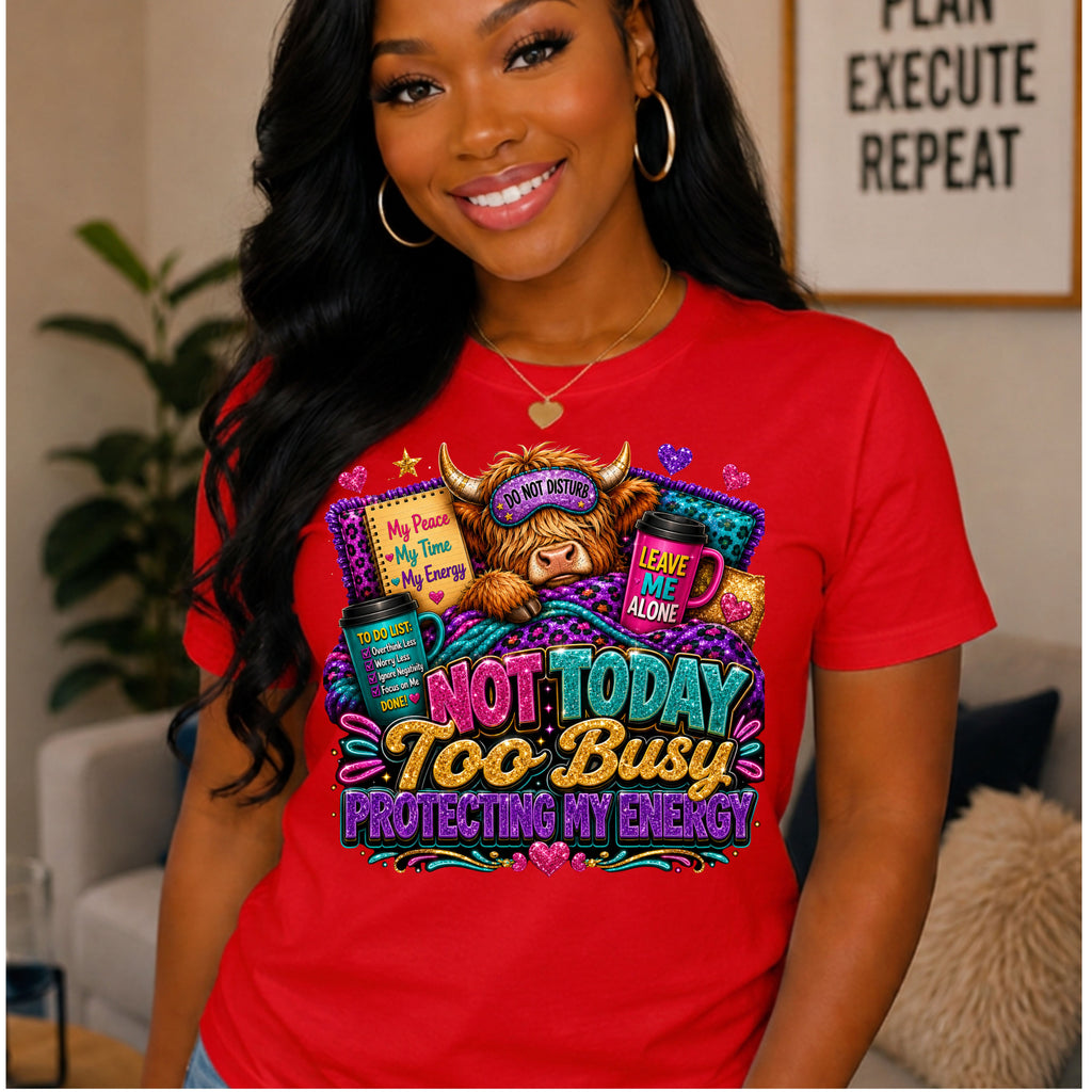 A smiling woman wearing a bright red graphic T-shirt in a cozy indoor setting. The shirt features a colorful cartoon illustration of a cow with a sleep mask and the text 'NOT TODAY TOO Busy PROTECTING MY ENERGY'.