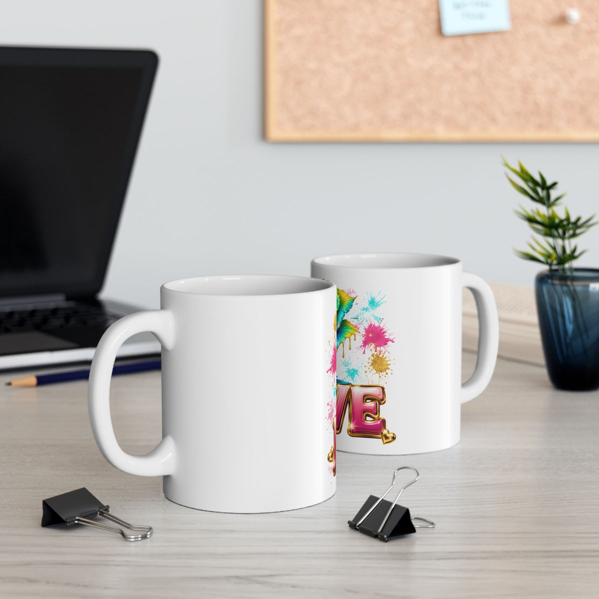 Two white ceramic mugs on a light wood desk in an office setting, with one mug showing a colorful graphic print and the other plain white. A laptop, binder clips, and a small plant are visible in the background.